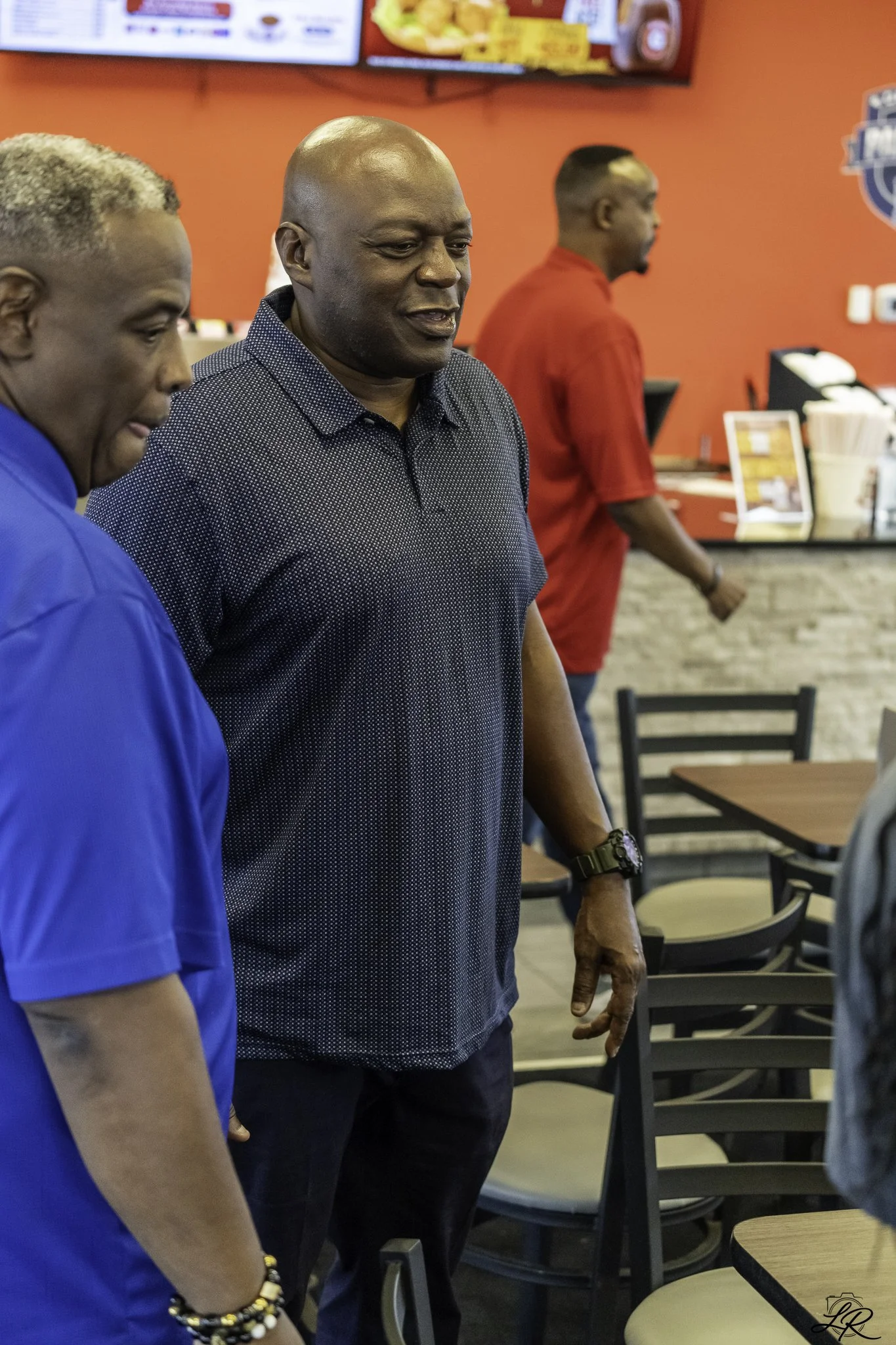 Three men standing in a casual restaurant or fast-food place with red walls. The man in the middle, who is bald and wearing a dark patterned shirt, appears to be talking. The other man, partially visible, has short, bleached hair and is wearing a blu
