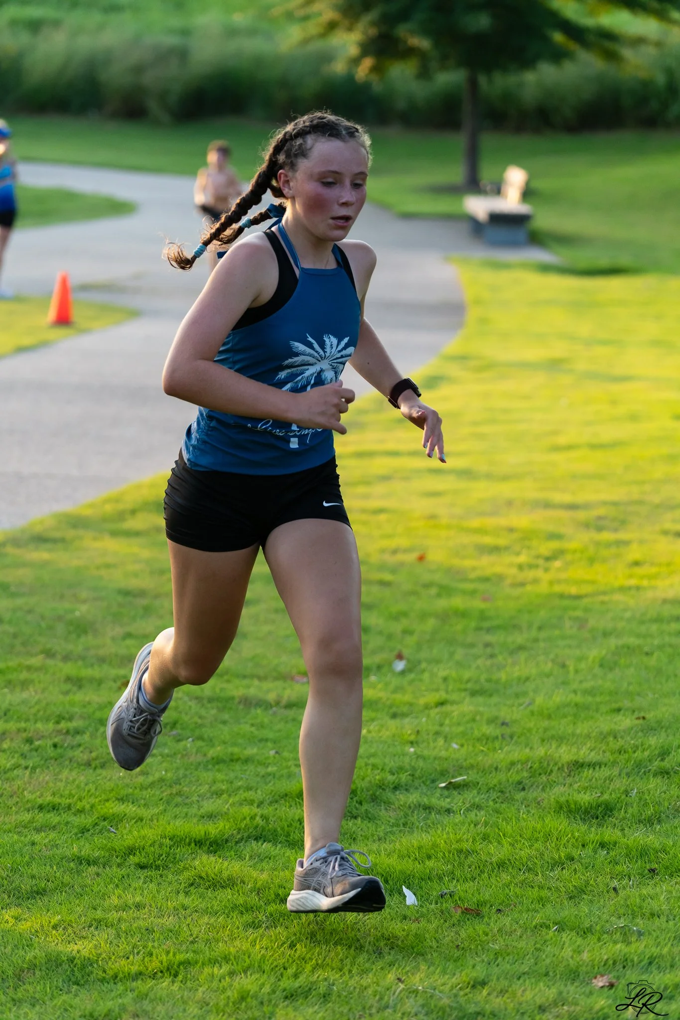 A young girl running outdoors on a grassy park path during daytime, wearing athletic clothes and sneakers, with other children visible in the background.
