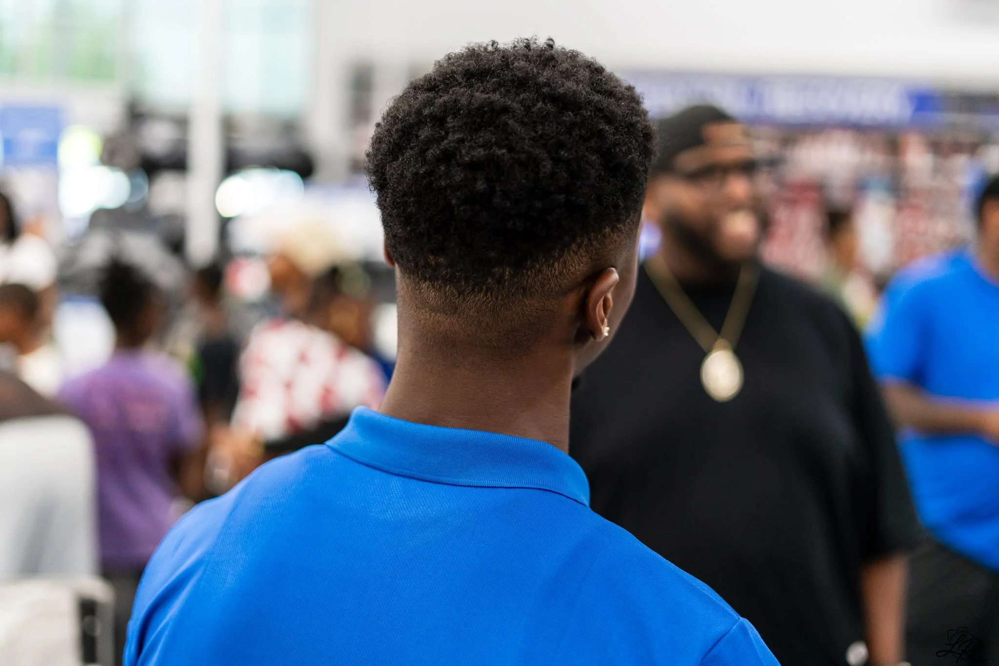 Back of a person with short curly hair wearing a blue shirt, in a busy indoor setting with people and bright lights in the background.
