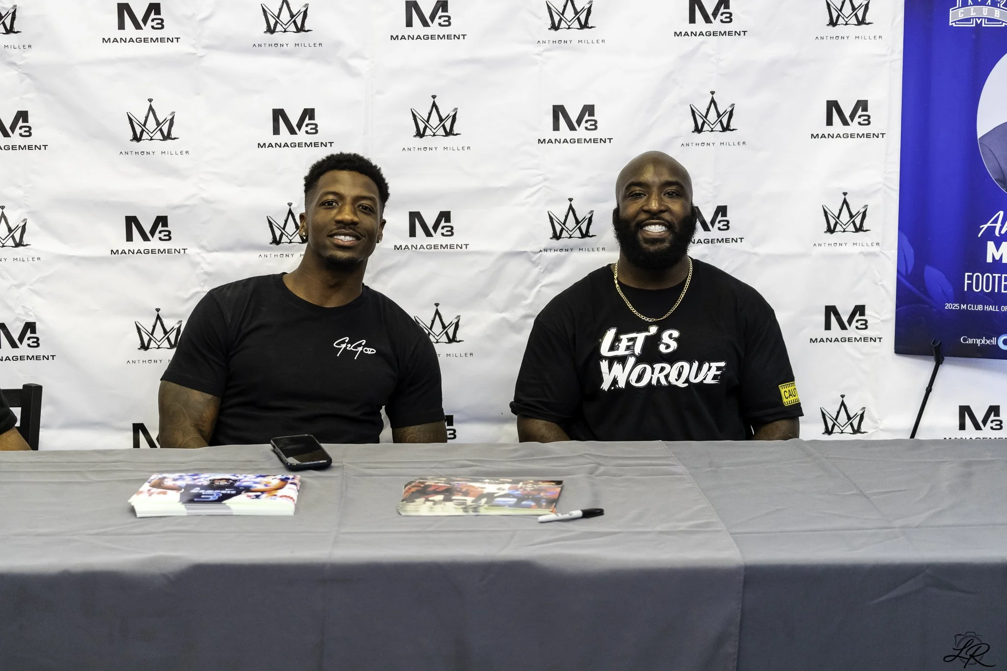 Two men sitting at a table during a signing event or press conference, with a backdrop featuring logos for Anthony Miller and M3 Management. One man has a black shirt with white text, and the other man has a black shirt with yellow text. There are ma