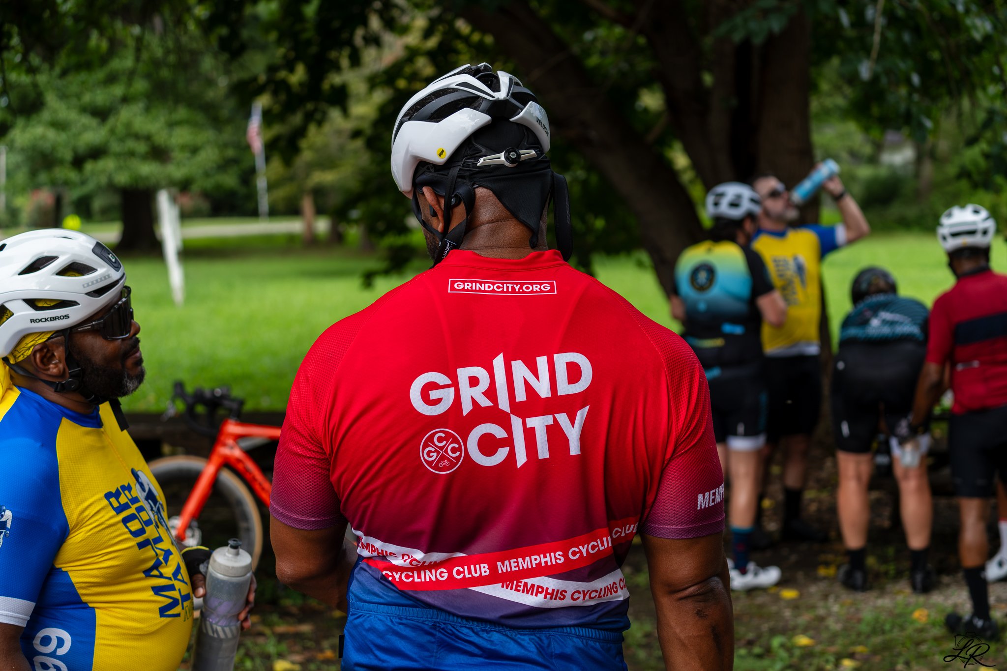 Cyclists gathered outdoors, some drinking water, with helmets and athletic gear, under trees in a park.