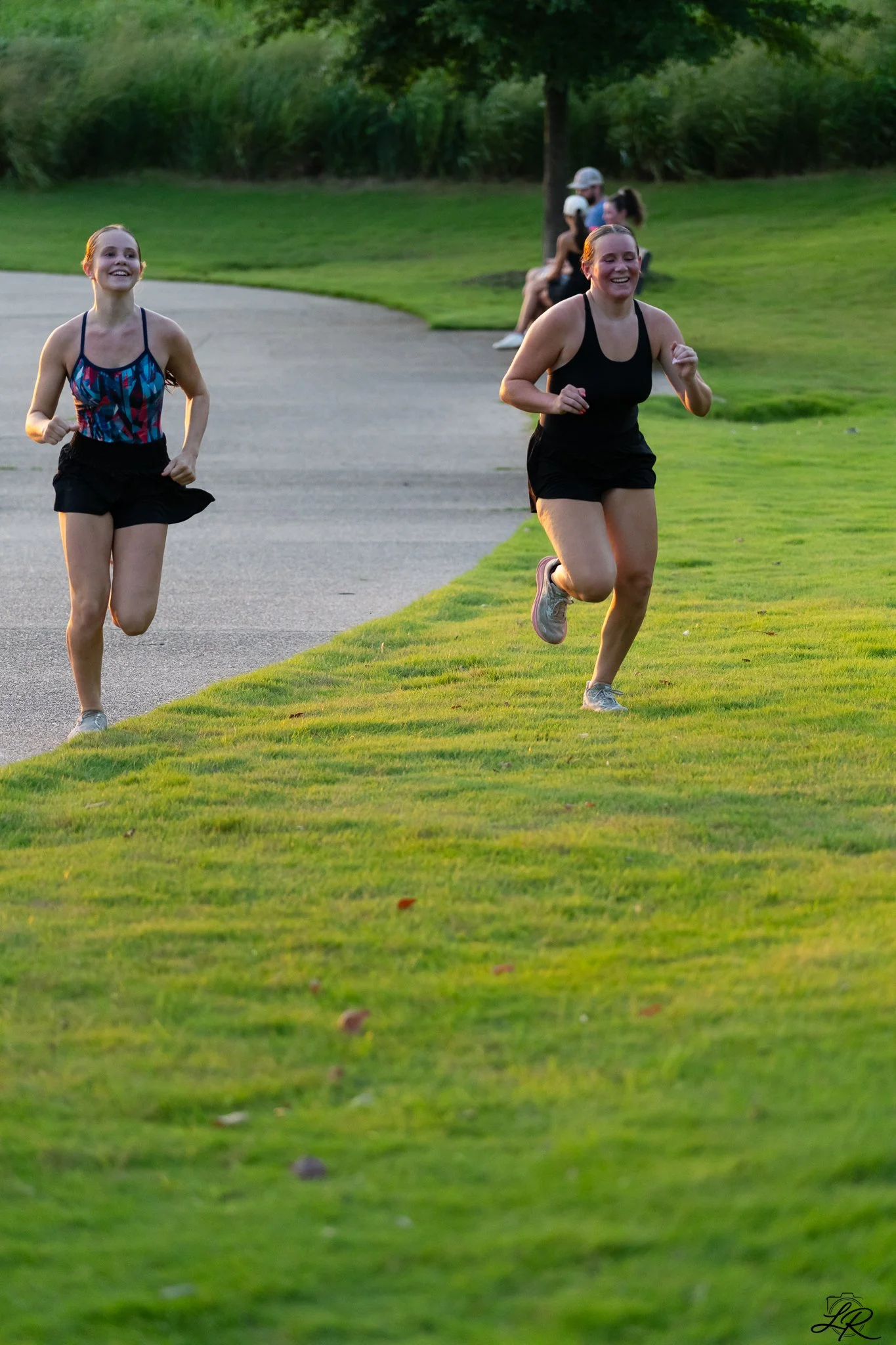 Two young women running on a grassy area in a park, with trees and benches in the background. They are smiling and appear to be enjoying themselves.