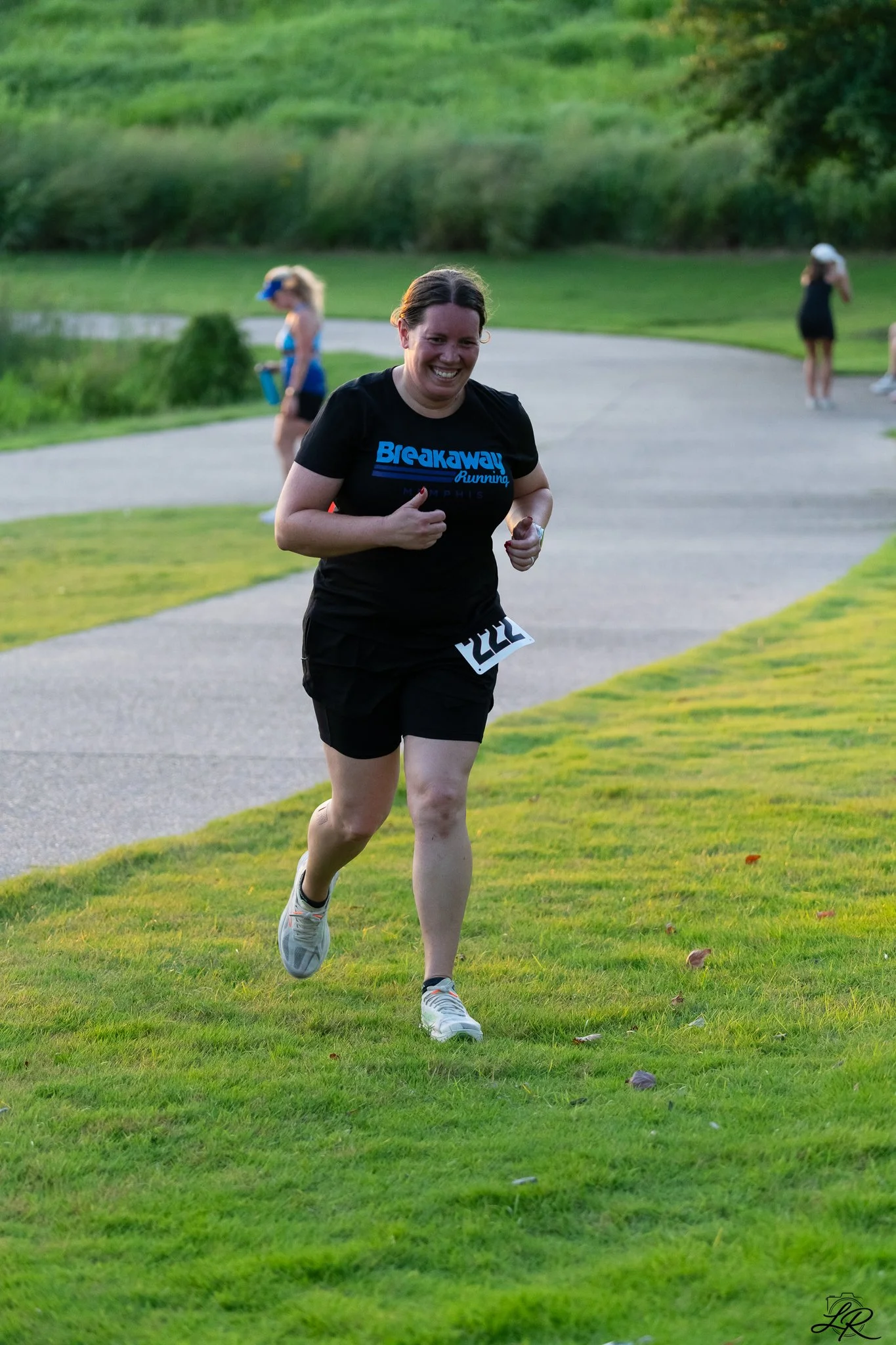 A woman running outdoors on a grassy path during daytime, wearing a black T-shirt with blue text, black shorts, and gray running shoes. She is smiling as she runs, with a park and other people in the background.