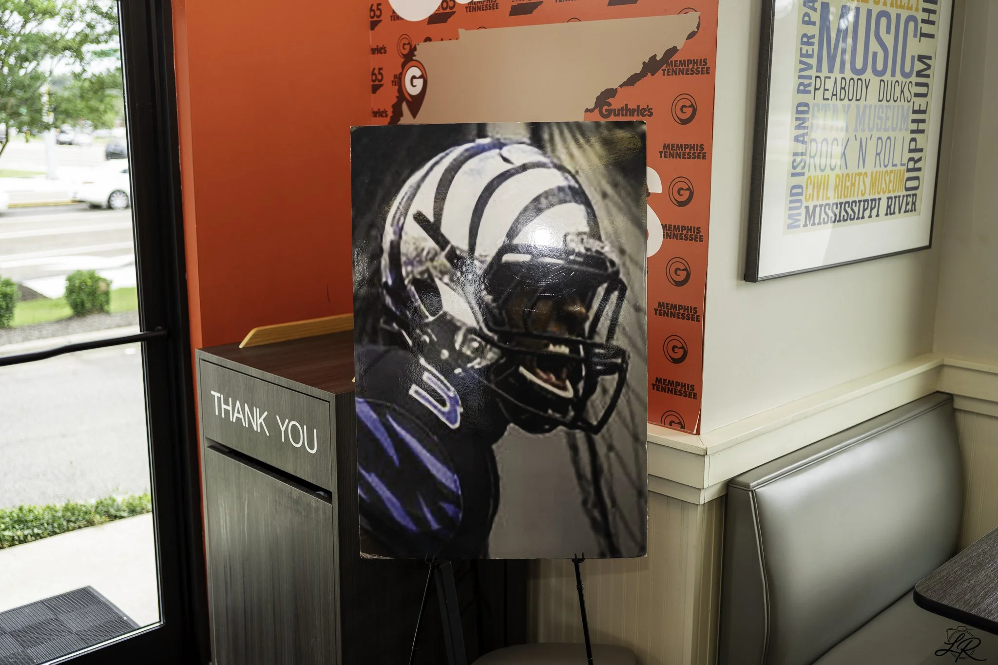 Indoor scene showing a poster of a football player in a helmet, a thank you sign on a gray box, a framed word cloud with locations and music genres, a red poster with a map of Tennessee, and part of a door with an exit mat outside.