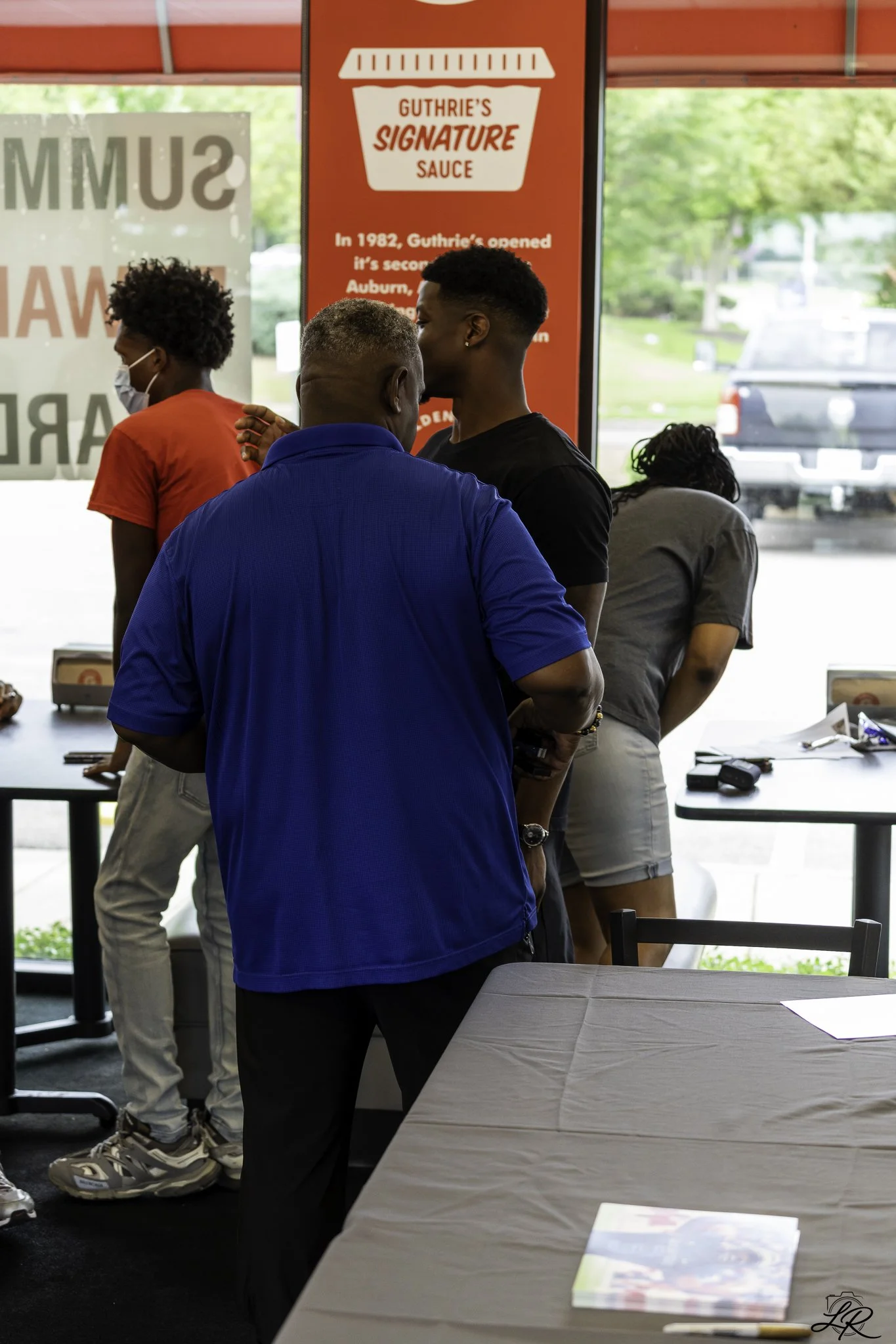 People standing indoors near a table with pamphlets, with a red sign behind them that reads "Guthrie's Signature Sauce." One person is wearing a face mask. Outside, there are trees and a truck visible through the window.