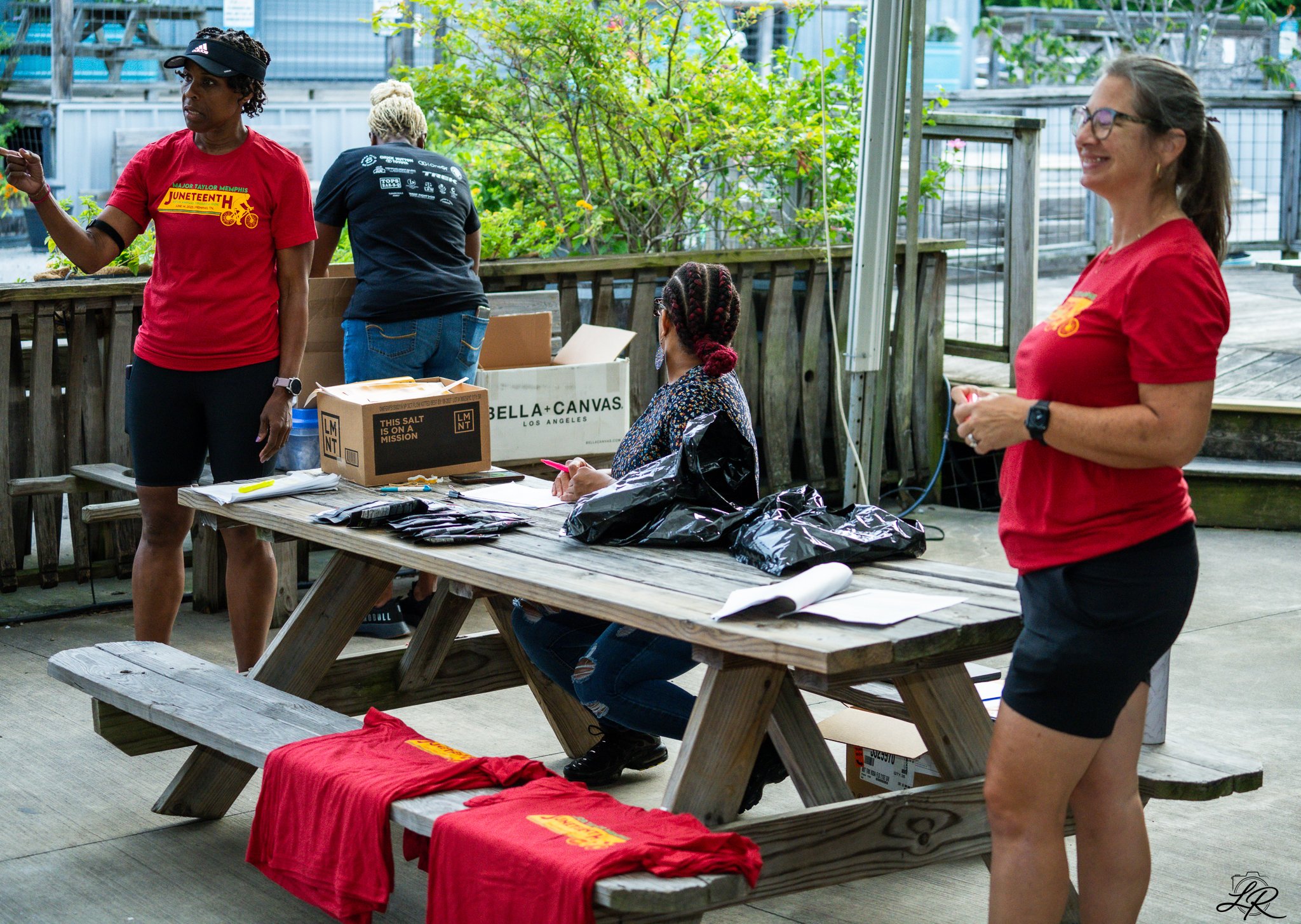Women at an outdoor table with T-shirts and boxes, some women are wearing red shirts, one woman is seated, others are standing, and a woman in the foreground is smiling and wearing glasses.