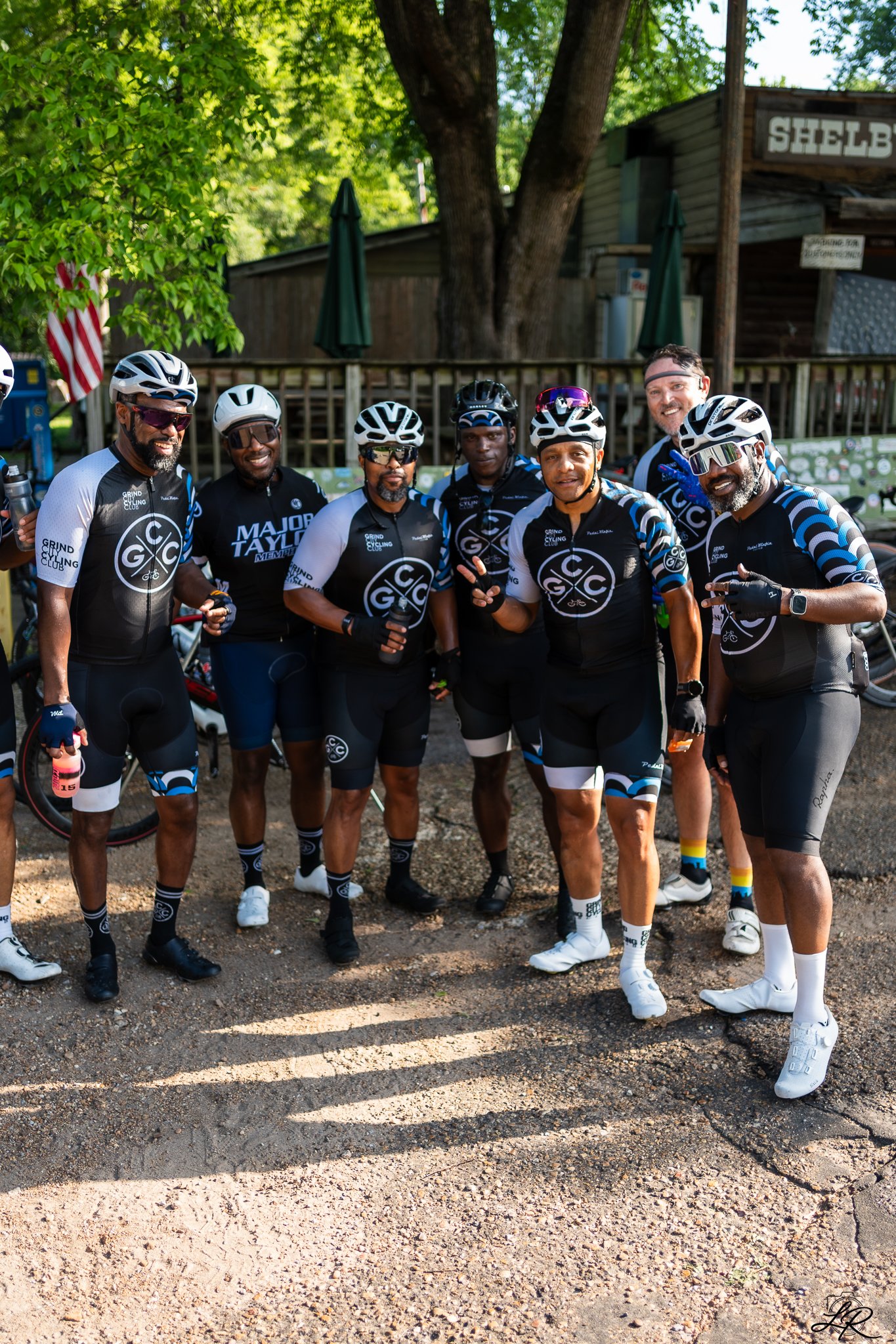 A group of eight diverse cyclists in cycling gear, helmets, and sunglasses pose outdoors for a photo on a sunny day. They are standing on a gravel surface with trees and a wooden building in the background.