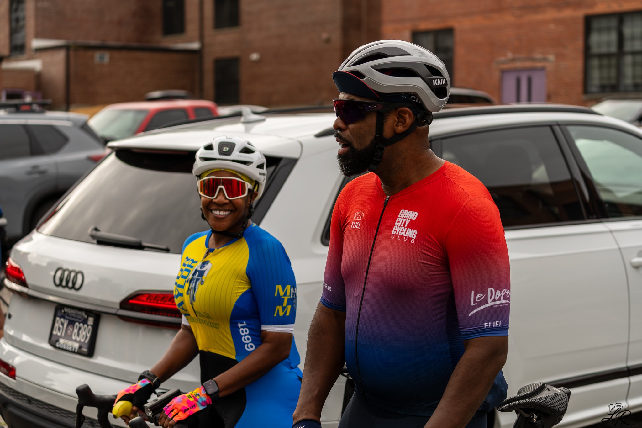 Two cyclists, a woman and a man, standing next to their bikes in a parking lot with brick buildings in the background. They are wearing helmets and cycling jerseys, and appear to be chatting.