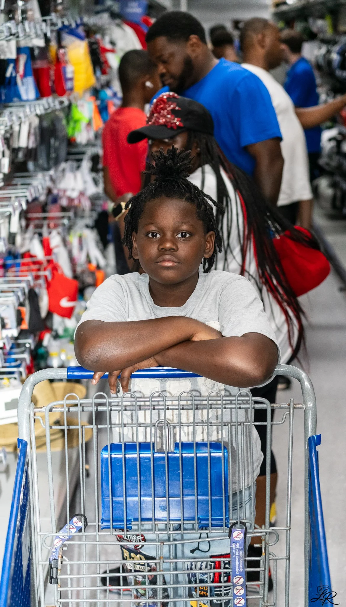 A young boy with dreadlocks sitting in a shopping cart at a store, with arms crossed on the cart's handlebar, looking directly at the camera. People are seen shopping in the background.