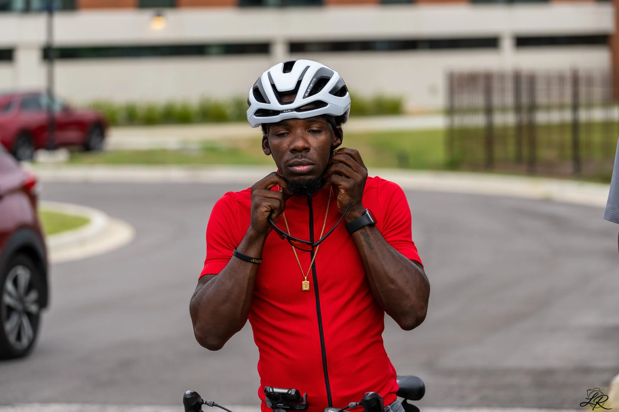 A man with dark skin wearing a white bicycle helmet and a red shirt, adjusting his helmet straps on a bicycle in a parking lot.