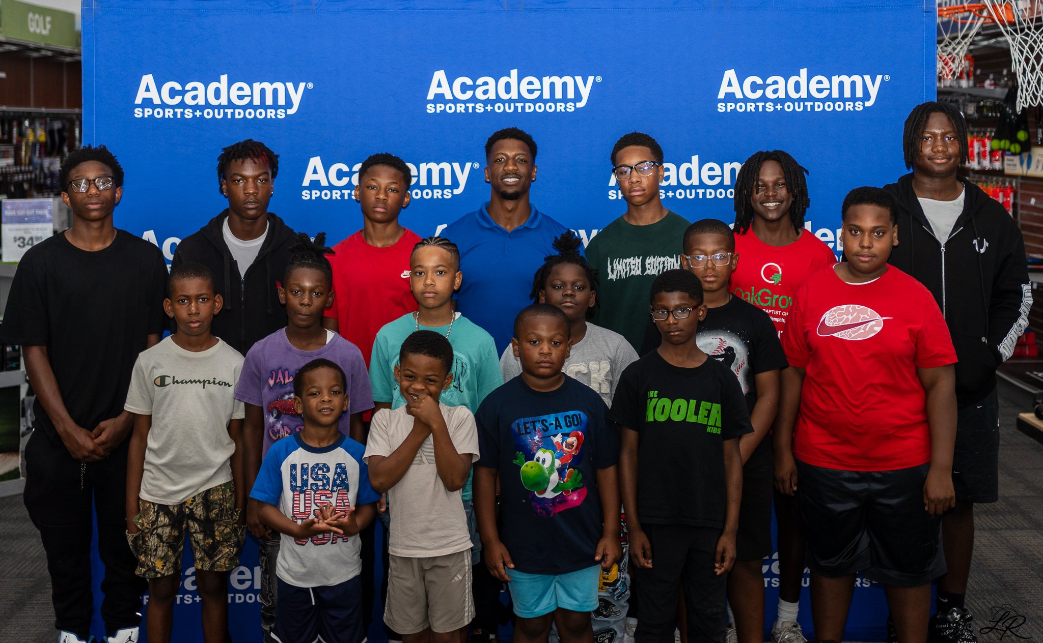 Group of children and young adults standing in front of a blue backdrop with 'Academy Sports+Outdoors' logos in a store.