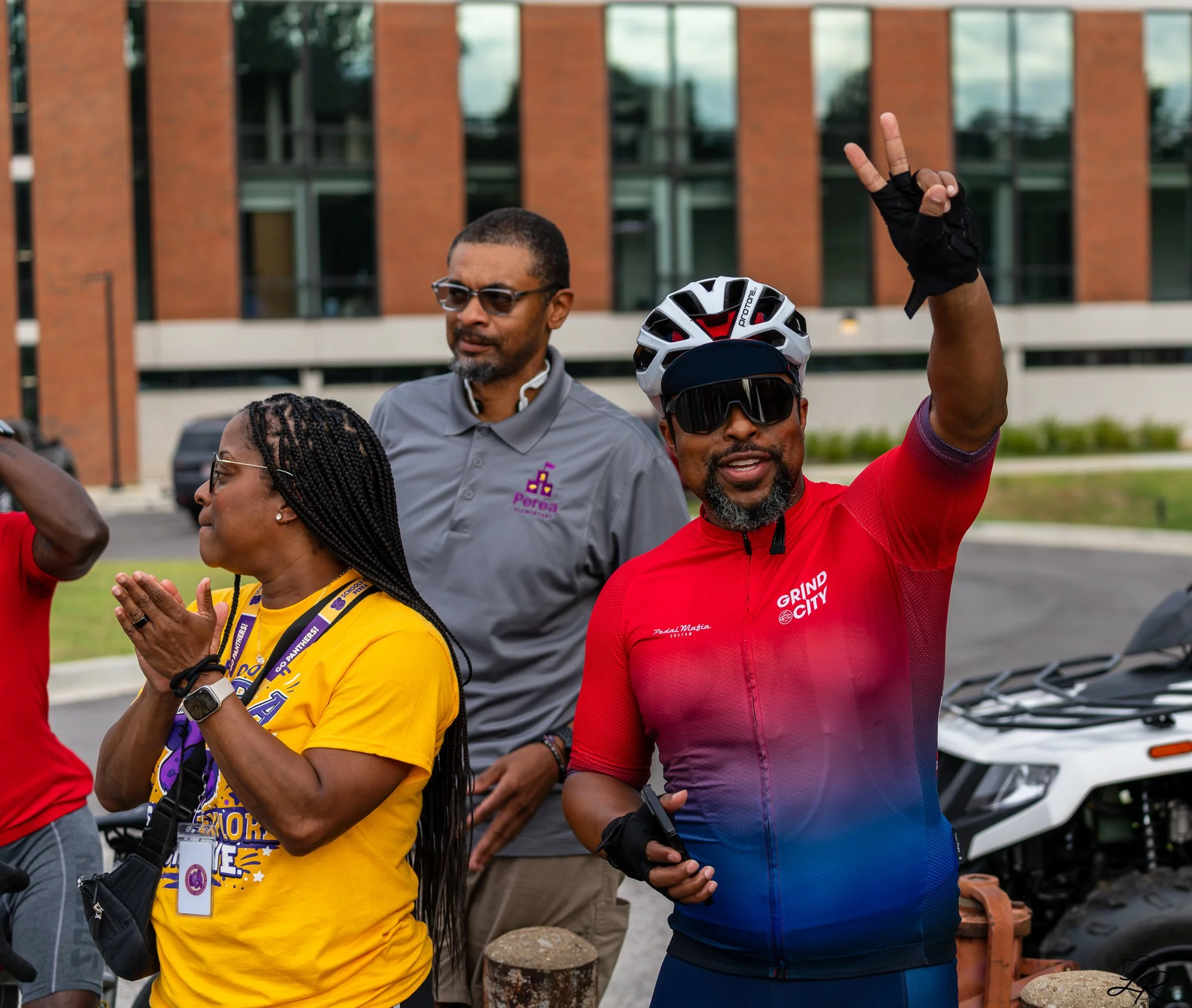 A diverse group of people outdoors, some wearing cycling gear and helmets, with one person making a peace sign and another praying, in front of a modern brick and glass building.