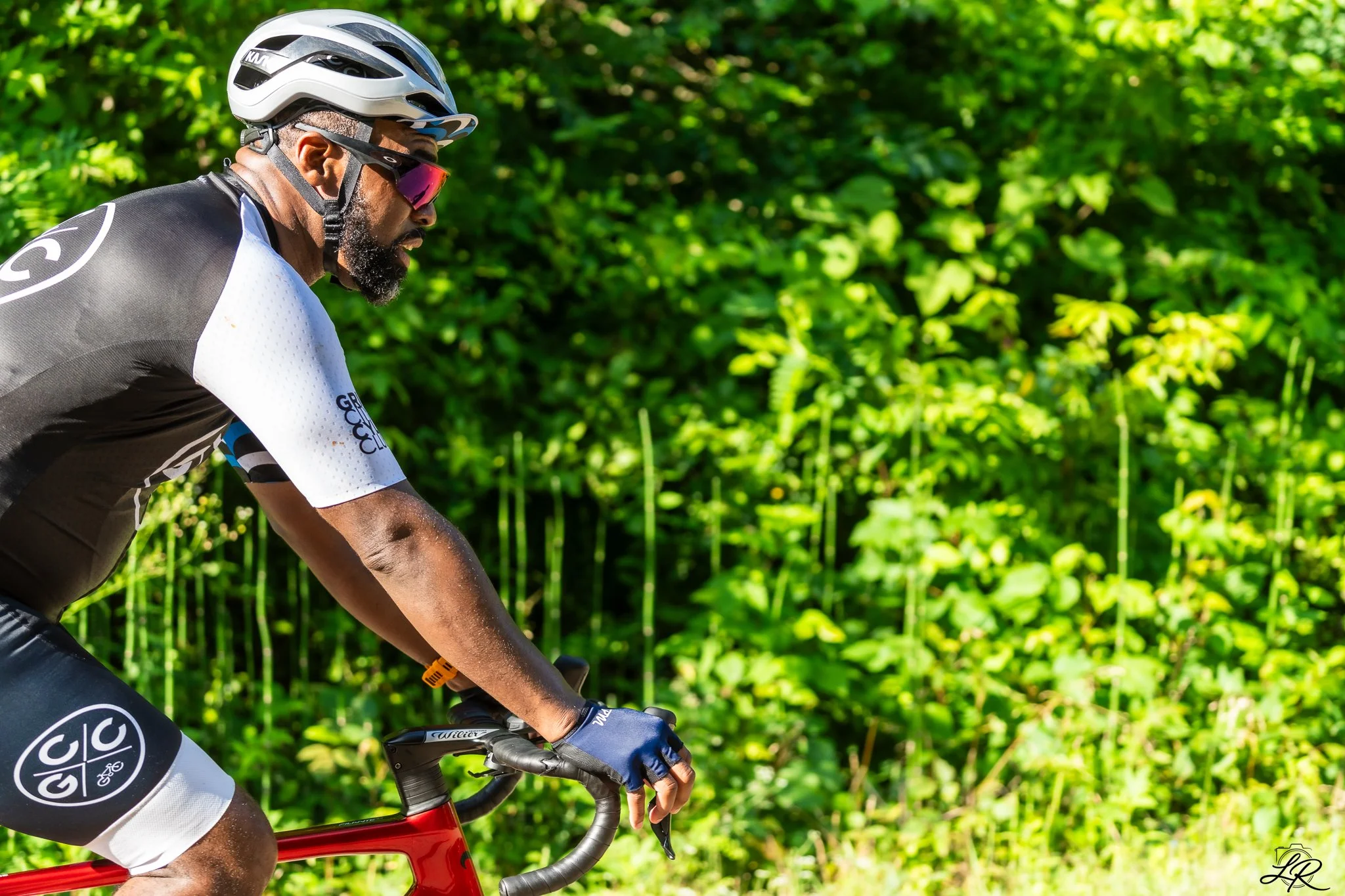 A man wearing a white cycling helmet, sunglasses, and a black and white cycling jersey riding a red bike through a green, lush area.