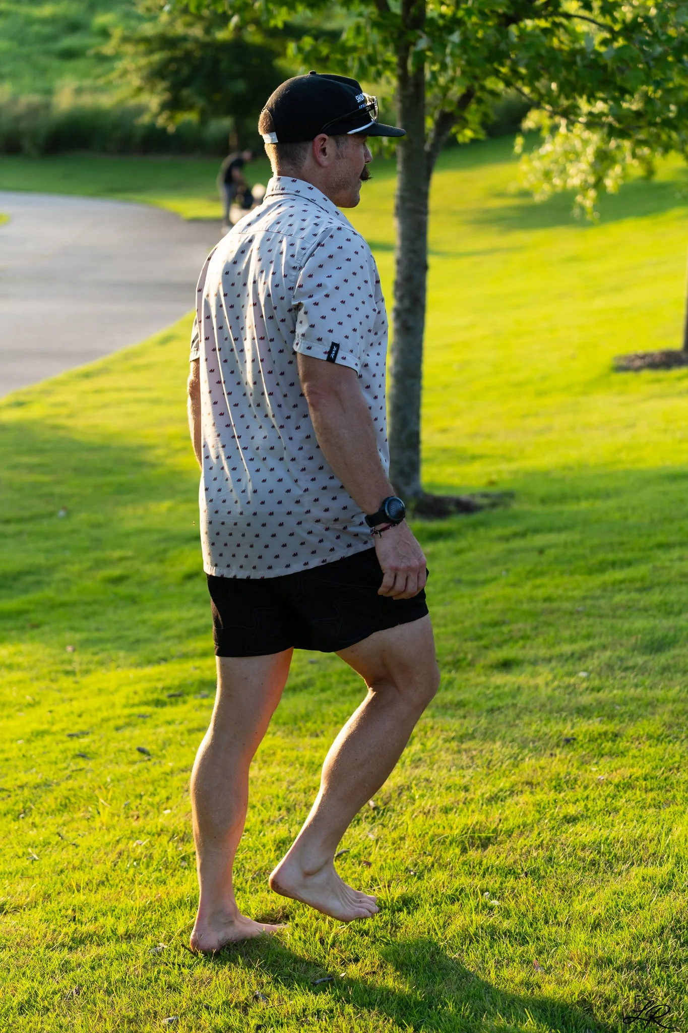 Man walking barefoot on grass in a park, wearing a patterned short-sleeve shirt and a black cap, with trees and a paved path in the background.