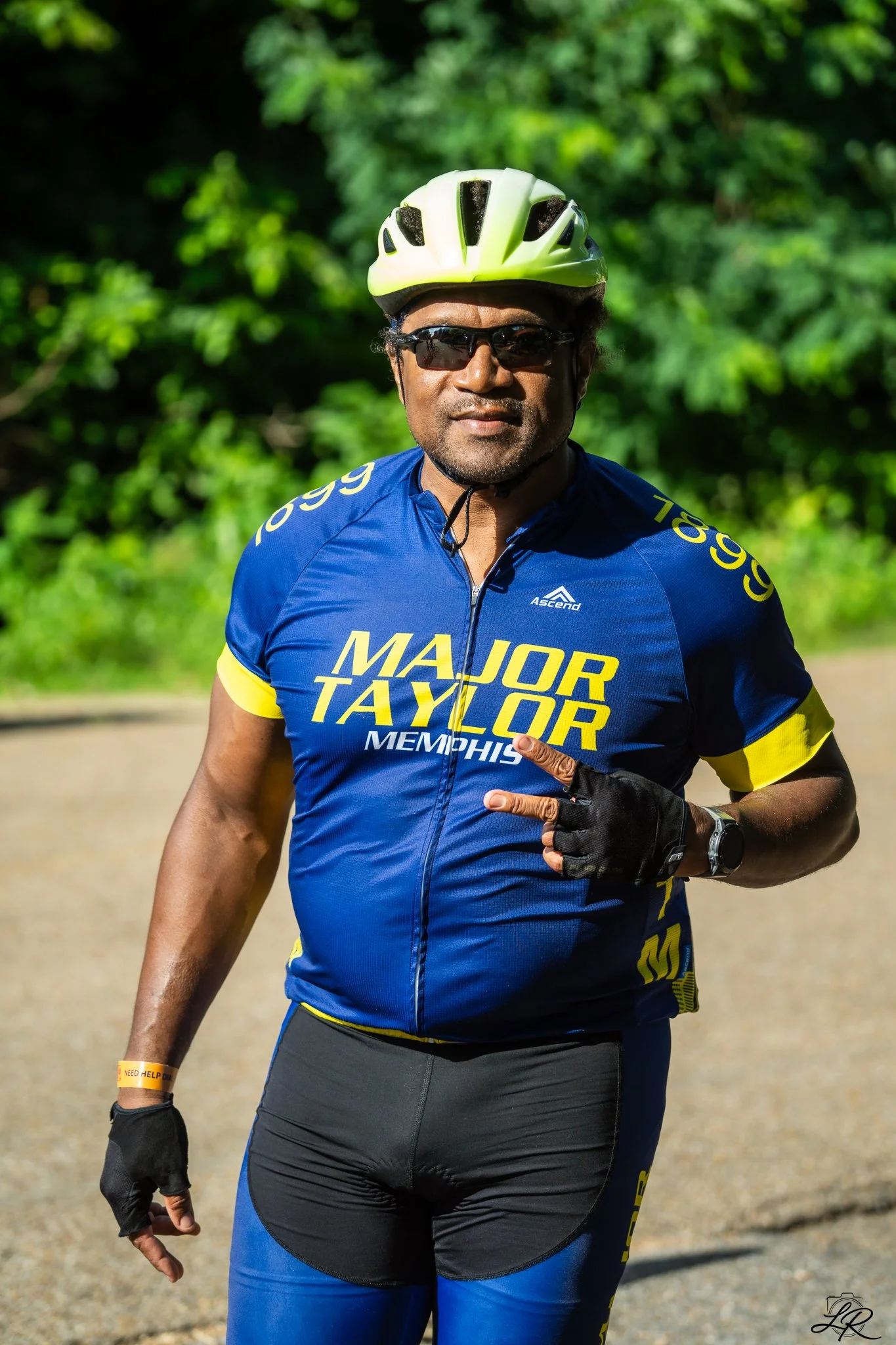 A man wearing a neon green bike helmet, sunglasses, and a blue Major Taylor Memphis cycling jersey, standing outdoors with greenery in the background.