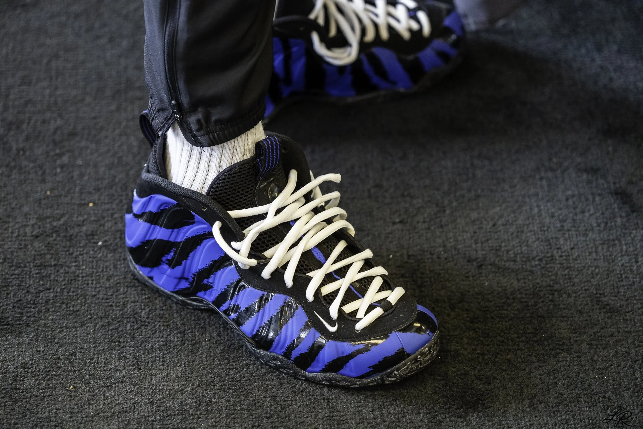 A close-up of a person wearing black and blue athletic shoes with white laces and black socks, standing on a dark carpeted floor, with part of their black pants visible.
