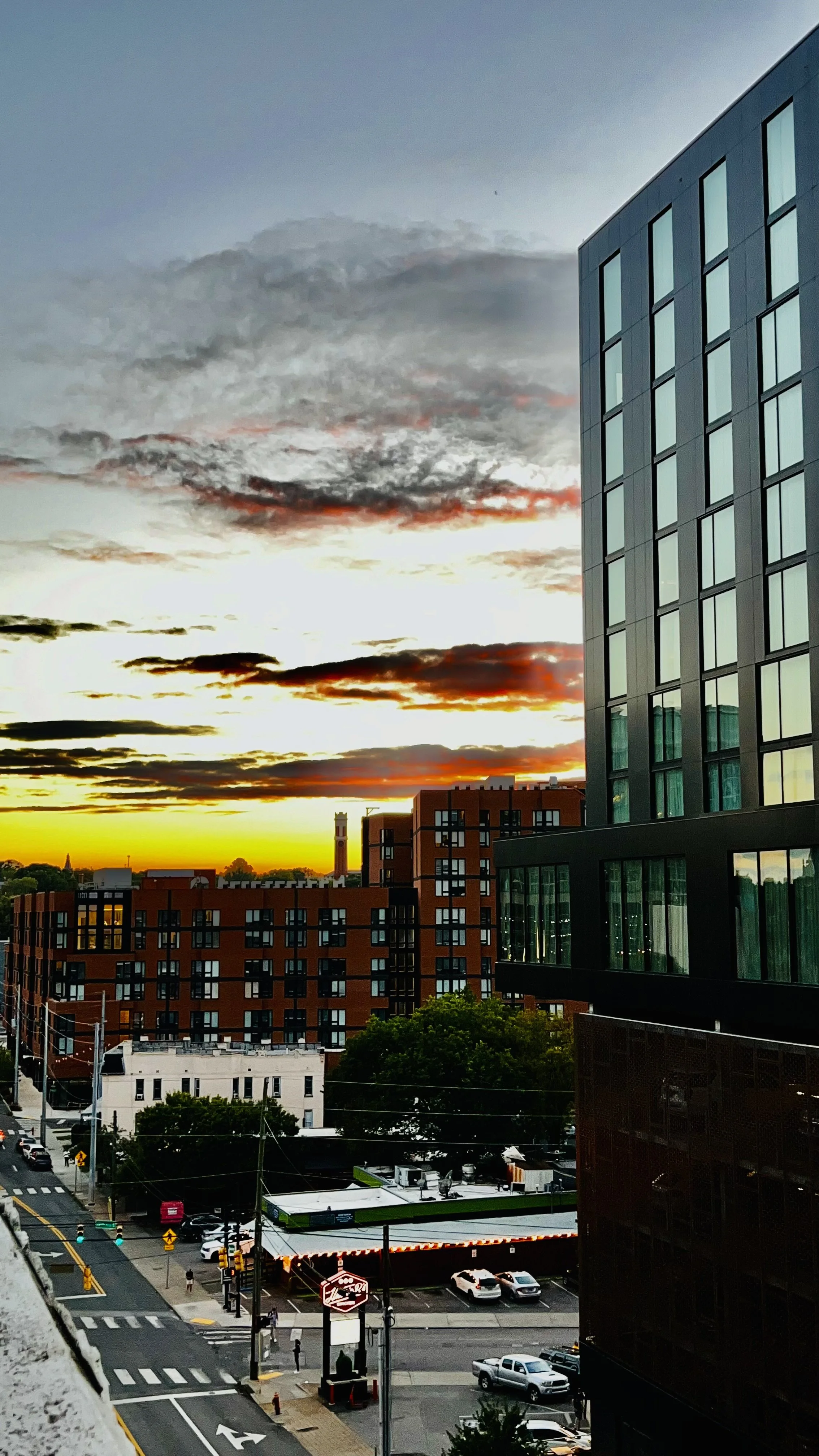 Cityscape during sunset with buildings, streets, cars, and a colorful sky with clouds.