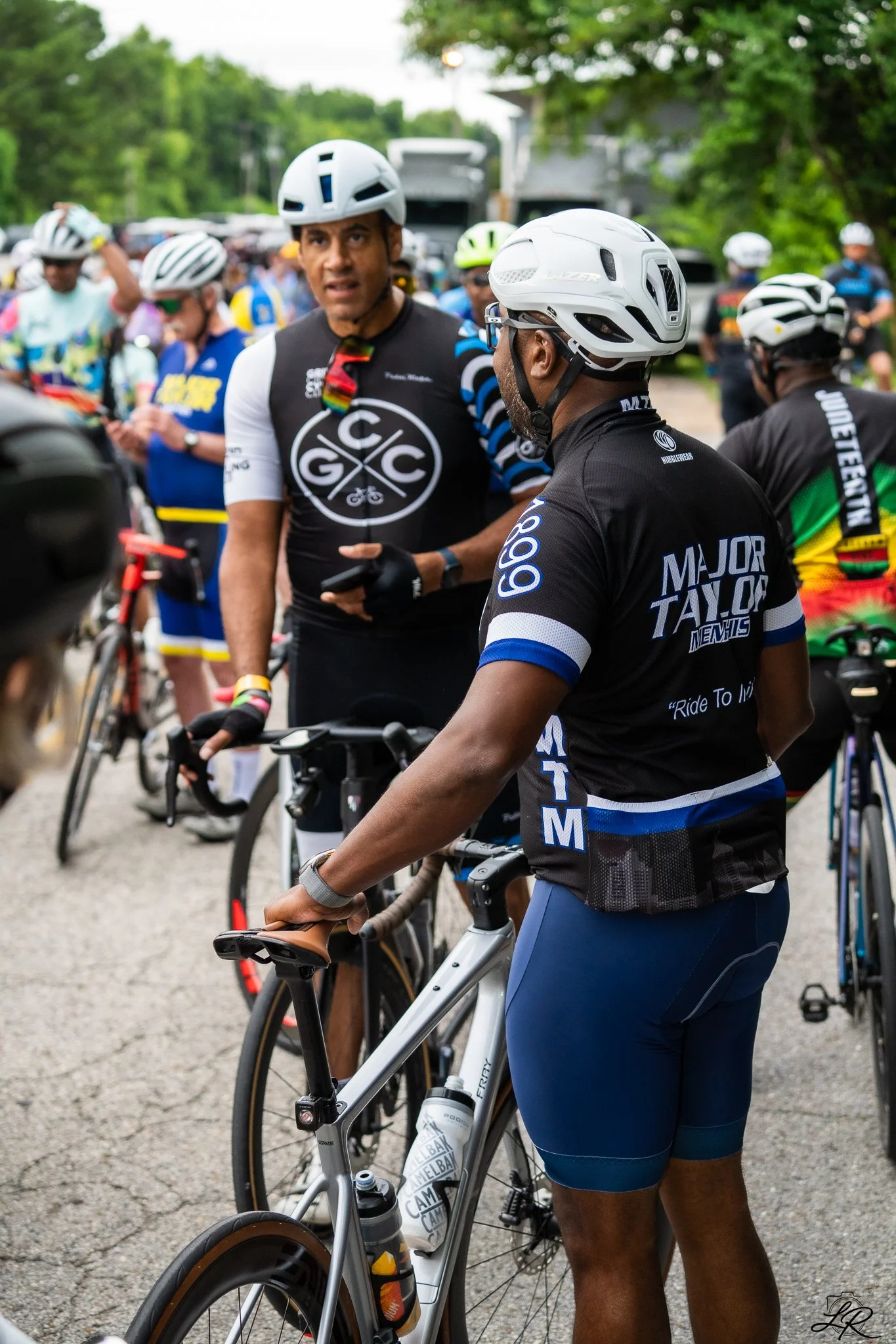A group of cyclists gathered outdoors, wearing helmets and athletic gear, preparing for a ride on a paved path surrounded by trees.