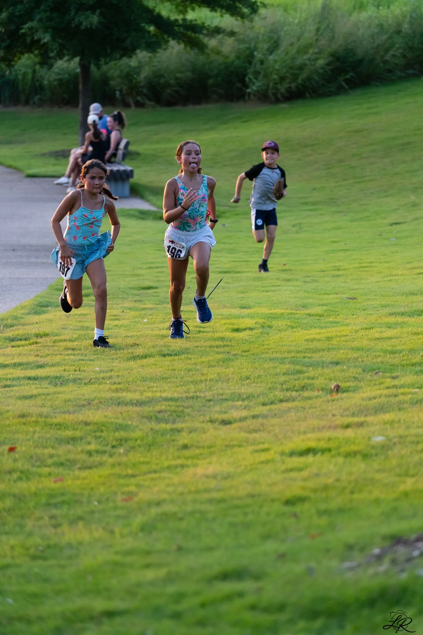 Three children running on a grassy field with trees and a park bench in the background, during what appears to be late afternoon or early evening.