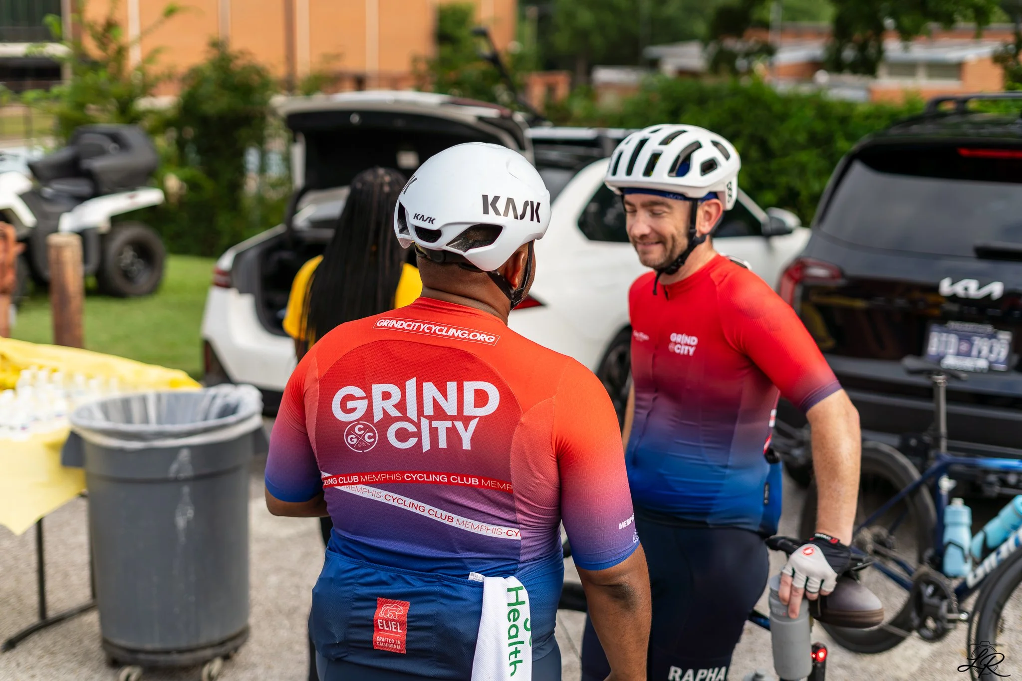 Two men wearing cycling helmets and red cycling jerseys with "GRIND CITY" printed on the back, standing outdoors near cars and trees, engaged in conversation at a biking event.