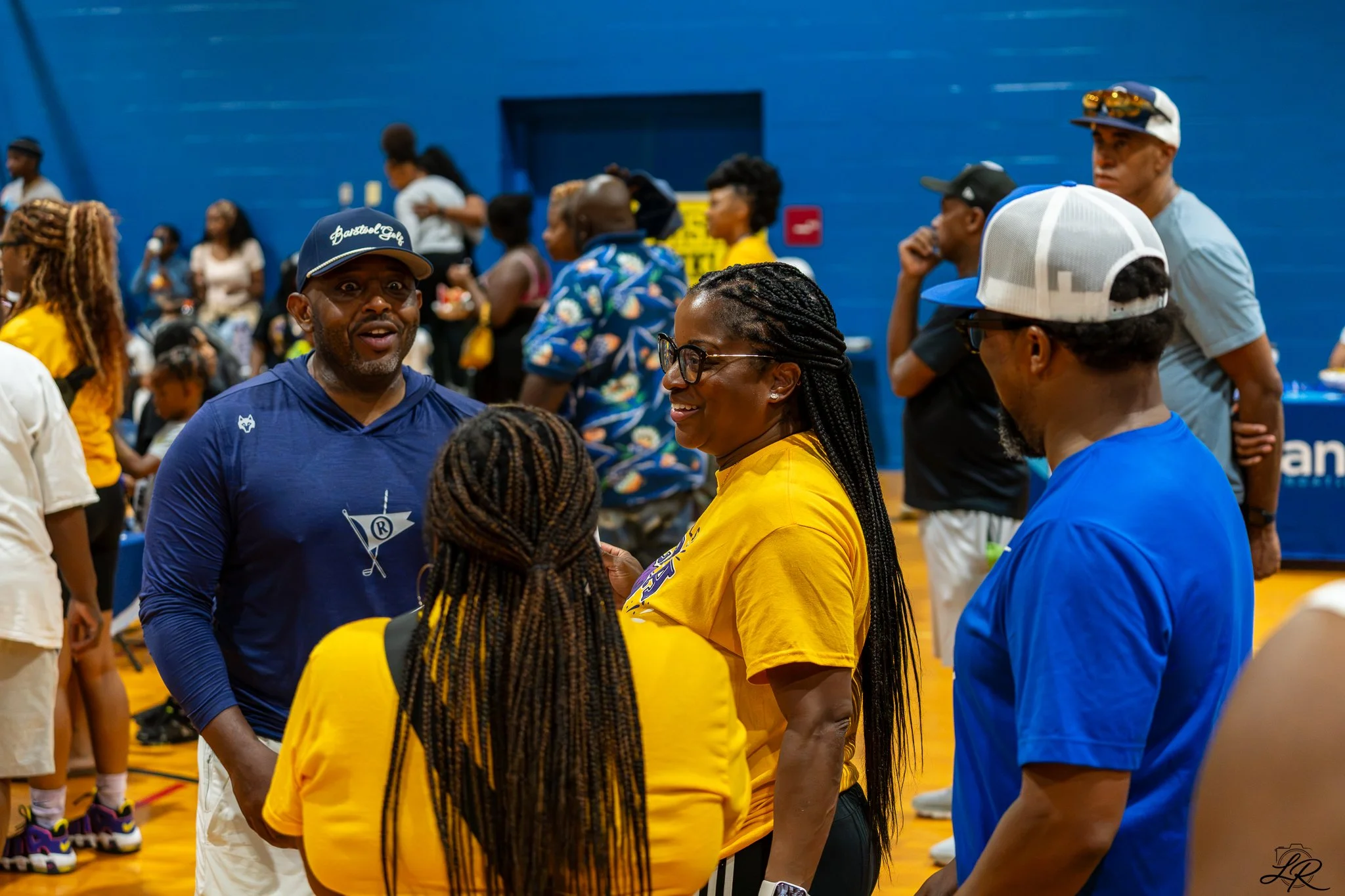 People socializing indoors with a blue wall in the background, some wearing colorful shirts and hats.