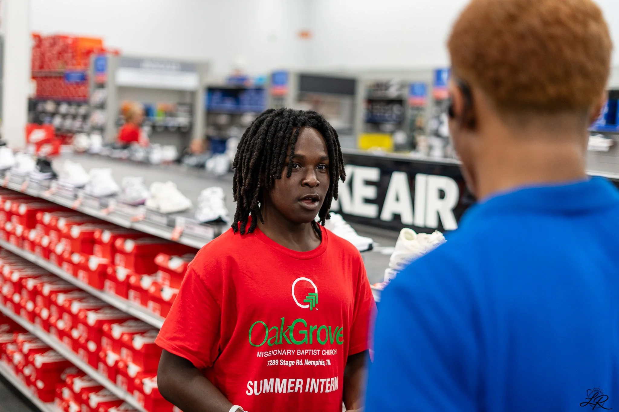 A young woman with dreadlocks wearing a red t-shirt with 'OakGrove' and 'MISSIONARY BAPTIST CHURCH' printed on it, talking to a young man with short curly hair in a blue shirt, in a store aisle with shoes on shelves.