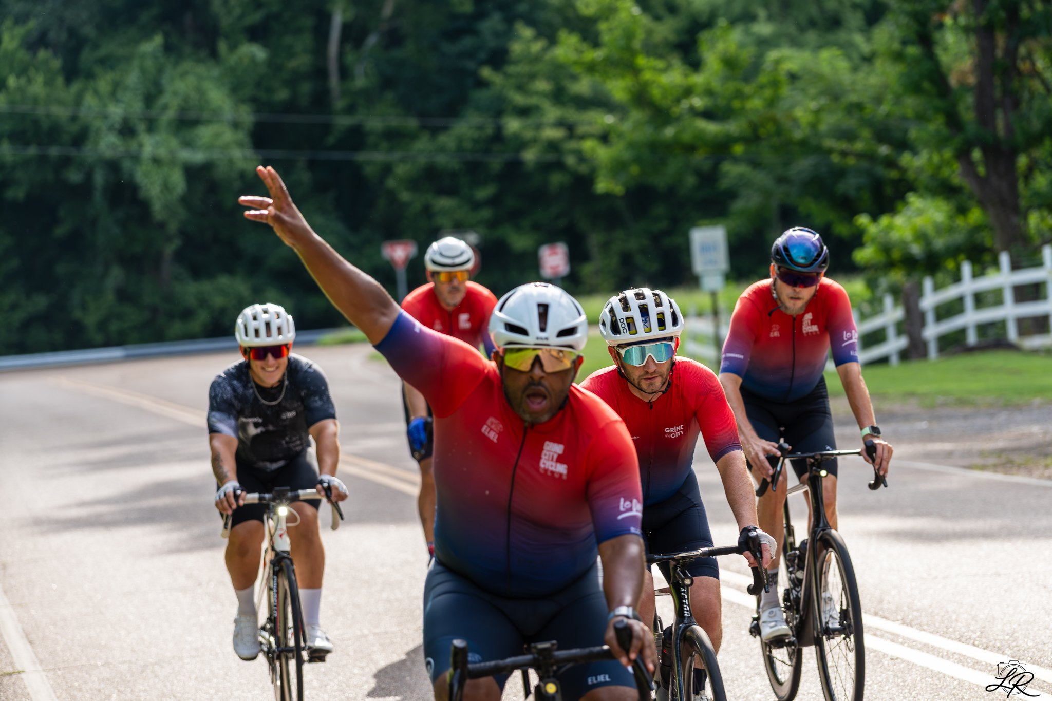 Group of five cyclists riding on a country road surrounded by green trees, with one cyclist in the foreground raising his arm and looking surprised or excited.