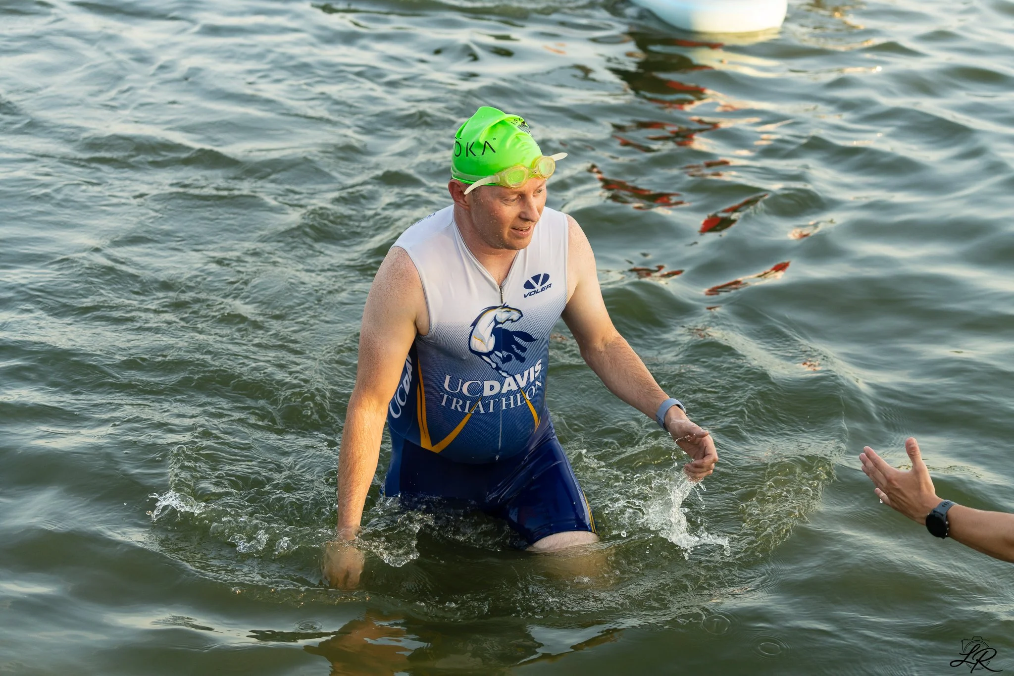 A male athlete wearing a green swim cap and goggles, dressed in a UC Davis Triathlon suit, is emerging from a body of water during a triathlon event.