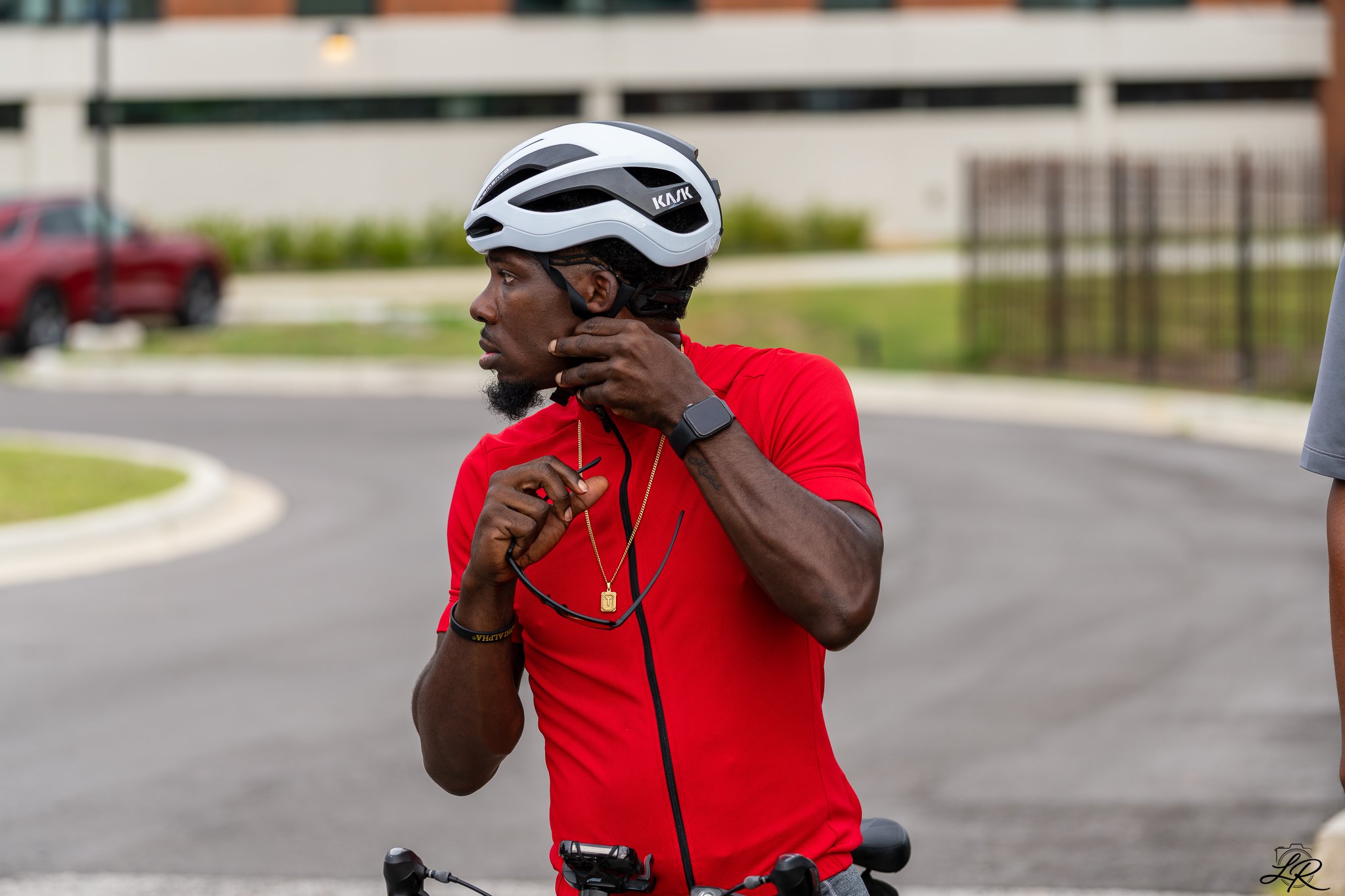 A man in a red shirt and a white bicycle helmet standing outdoors near a bicycle, adjusting his helmet strap with a focused expression.