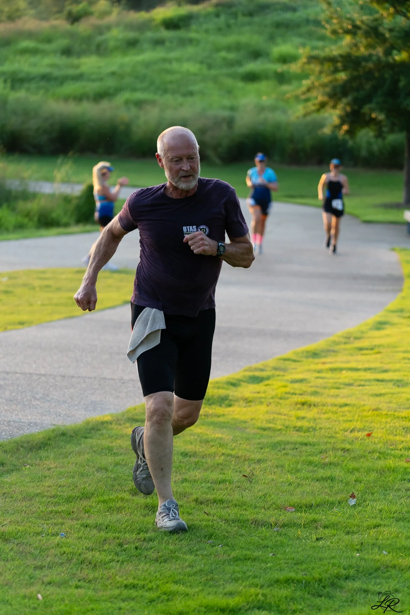 An elderly man running along a grassy park path with three women in athletic clothing running behind him. The park has lush green trees and a hill in the background, bathed in warm sunlight.