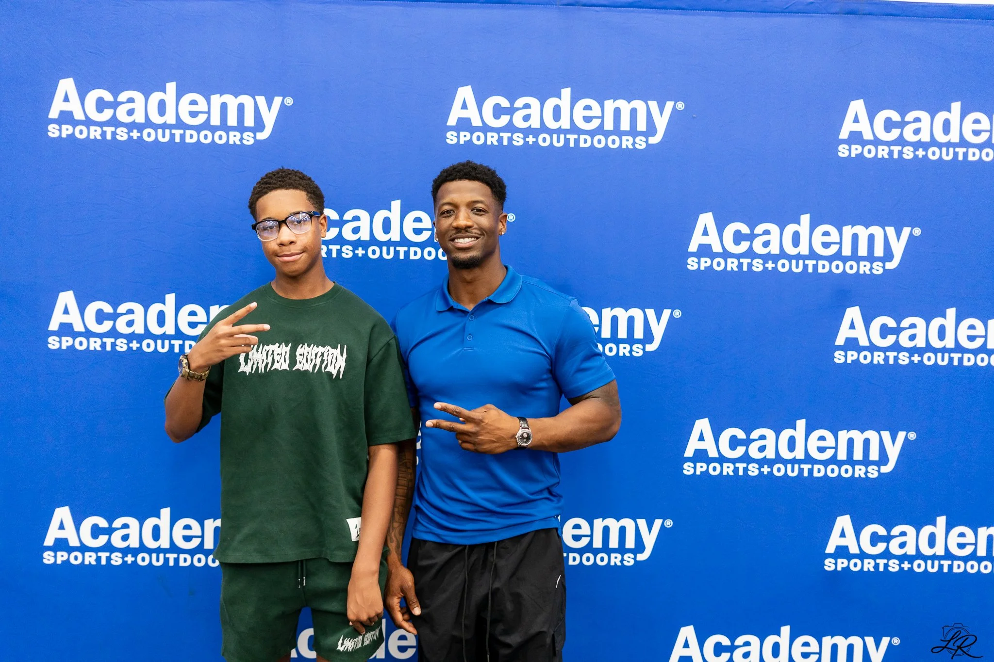Two young men standing in front of a blue backdrop with white text that reads "Academy Sports + Outdoors". One is wearing glasses and a green shirt, and the other is wearing a blue polo shirt, both making peace signs with their hands.