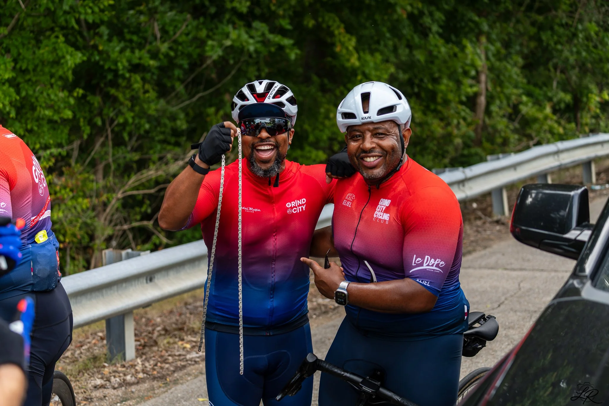Two men in red cycling jerseys and white bike helmets smiling and posing together outdoors, with greenery in the background.