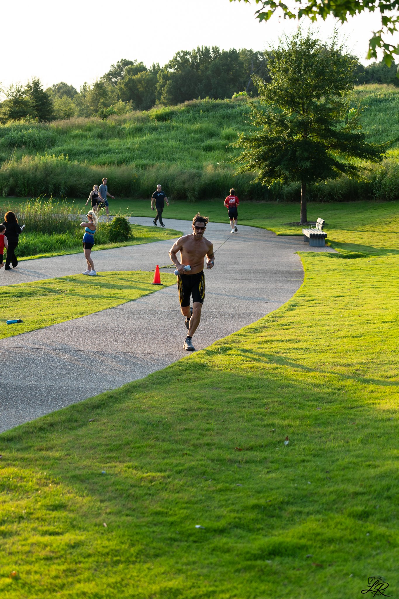 A park scene during sunset with a winding paved path, a shirtless man running, other people walking or stretching, lush green grass, a large tree, and a bench, with trees and hills in the background.