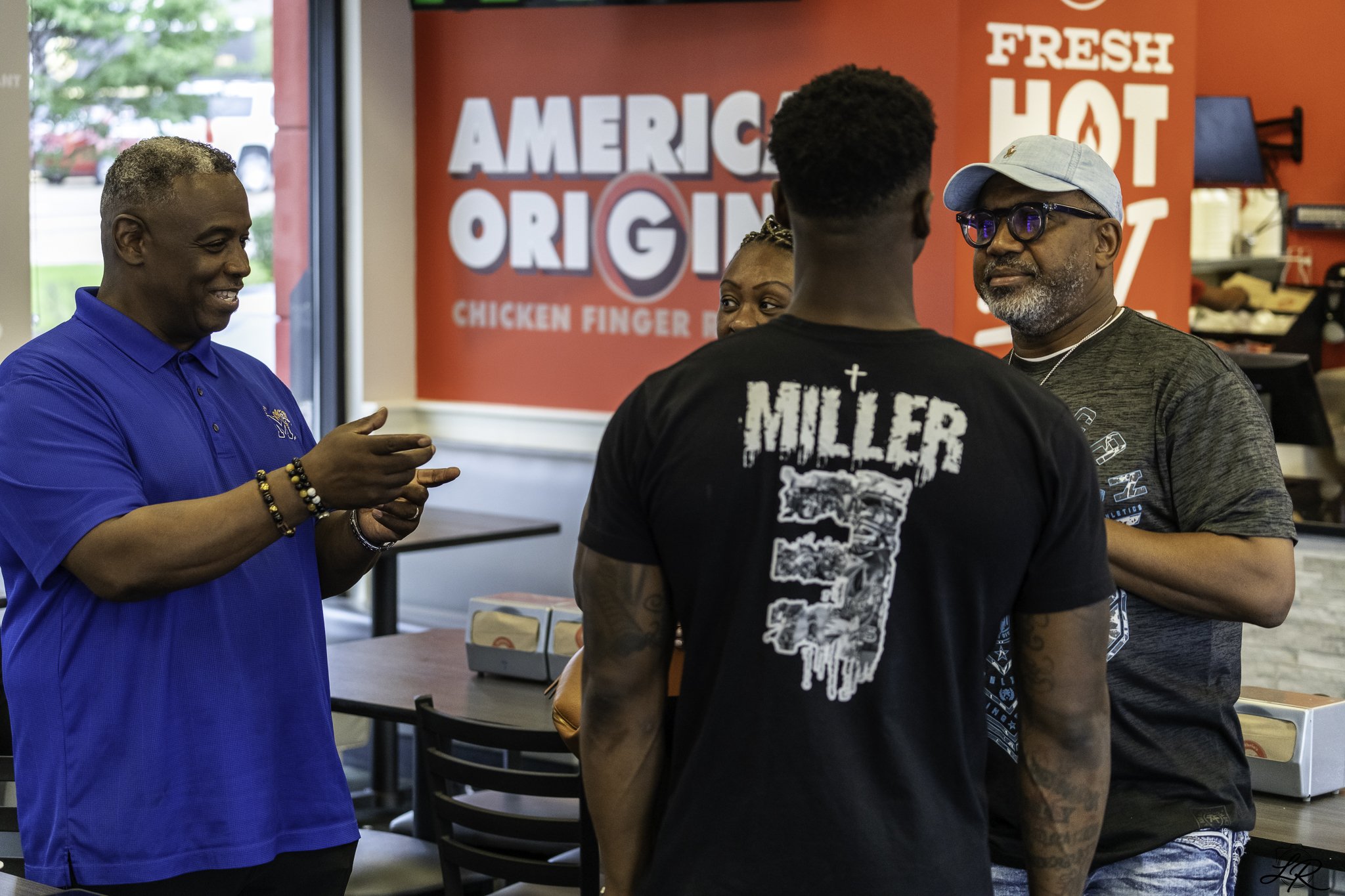 Four people engaged in conversation inside a fast food restaurant with a red wall that has the words "American Original" and "Fresh Hot" visible in the background.