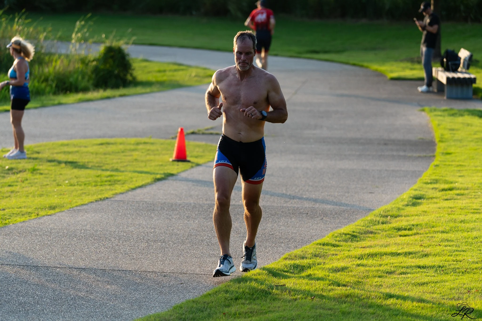 A shirtless man running on a paved path in a park during the daytime, with other people walking and sitting on benches in the background.