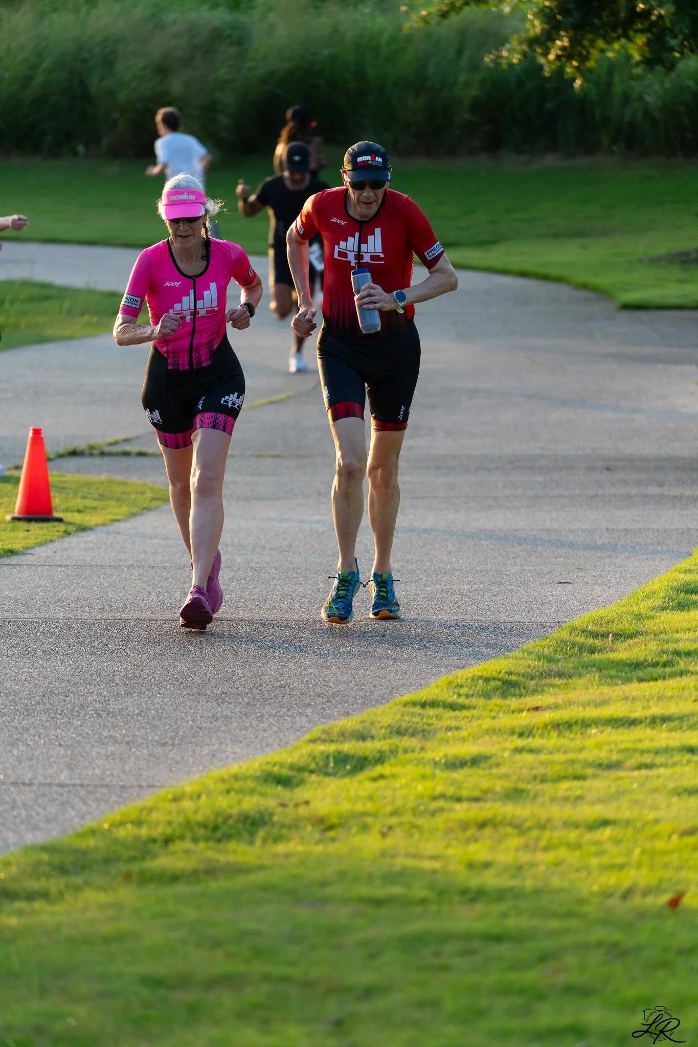 Two middle-aged runners, a woman and a man, participating in a race or training session outdoors on a path surrounded by green grass and trees. The woman is wearing a pink athletic outfit and a pink visor, and the man is in a red athletic shirt and b