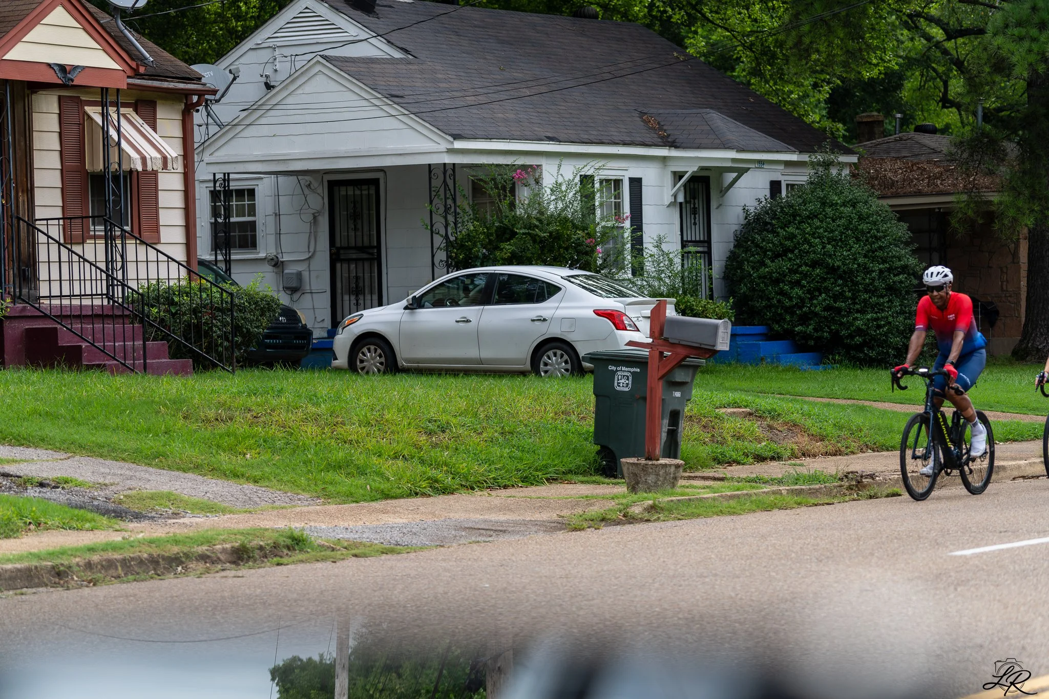 A residential street with a white house, parked cars, a mailbox, greenery, and a cyclist riding on the sidewalk.