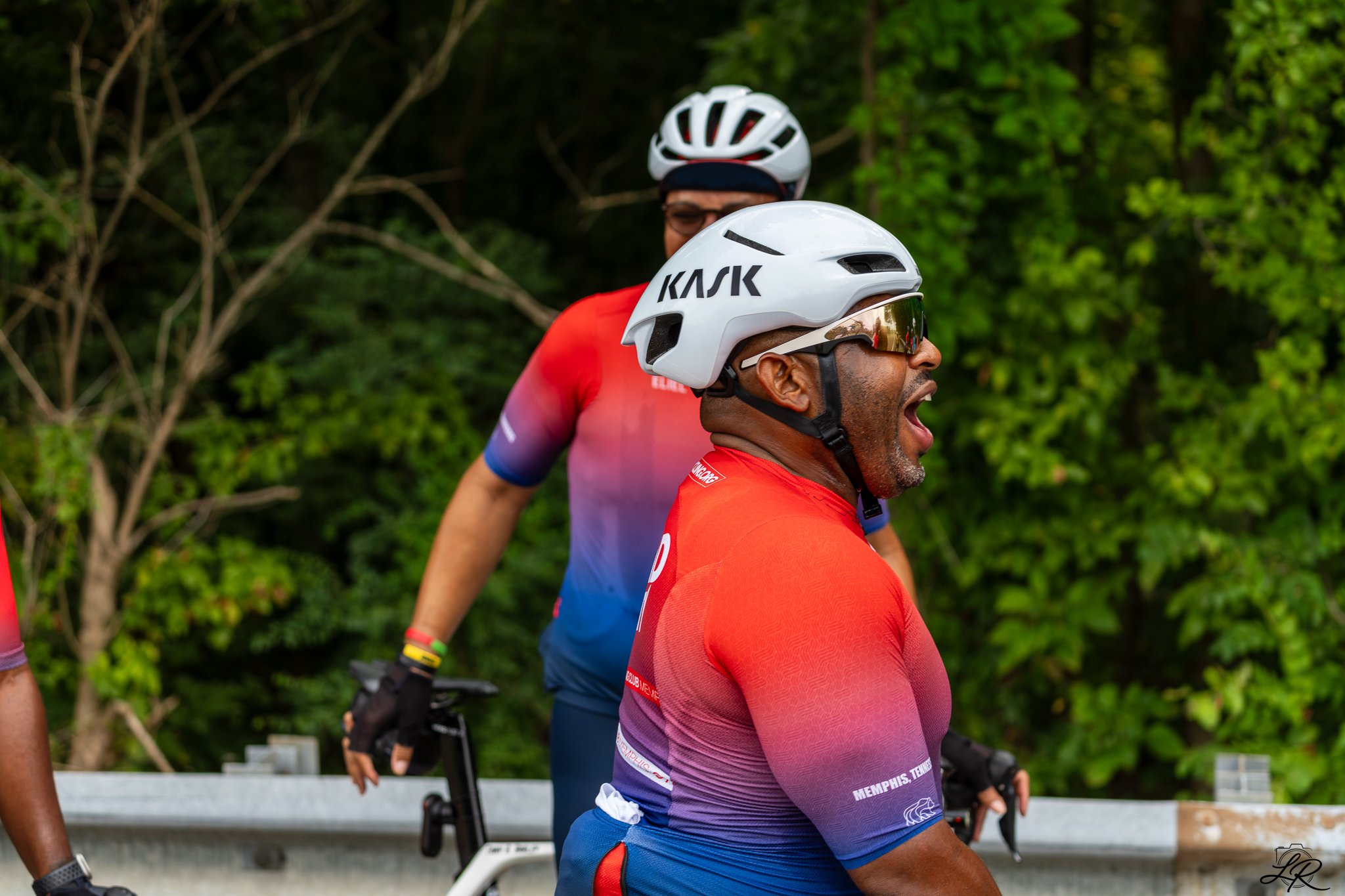 Two cyclists wearing helmets, one in the foreground with a white helmet and sunglasses, the other in the background with a white and black helmet, both wearing red and colorful cycling jerseys, standing outdoors with green trees in the background.