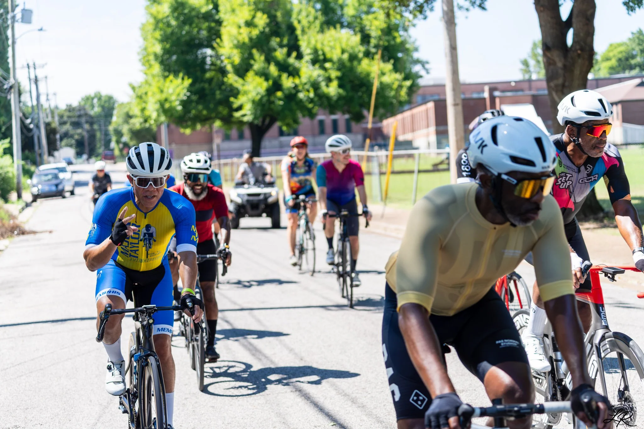 Cyclists riding together in a group on a sunny street with green trees and buildings in the background.