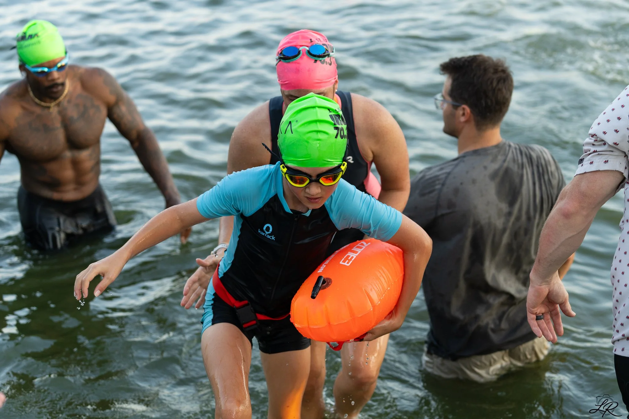Swimmers wearing colorful swim caps, goggles, and wetsuits in a body of water, with a person holding an orange rescue buoy.