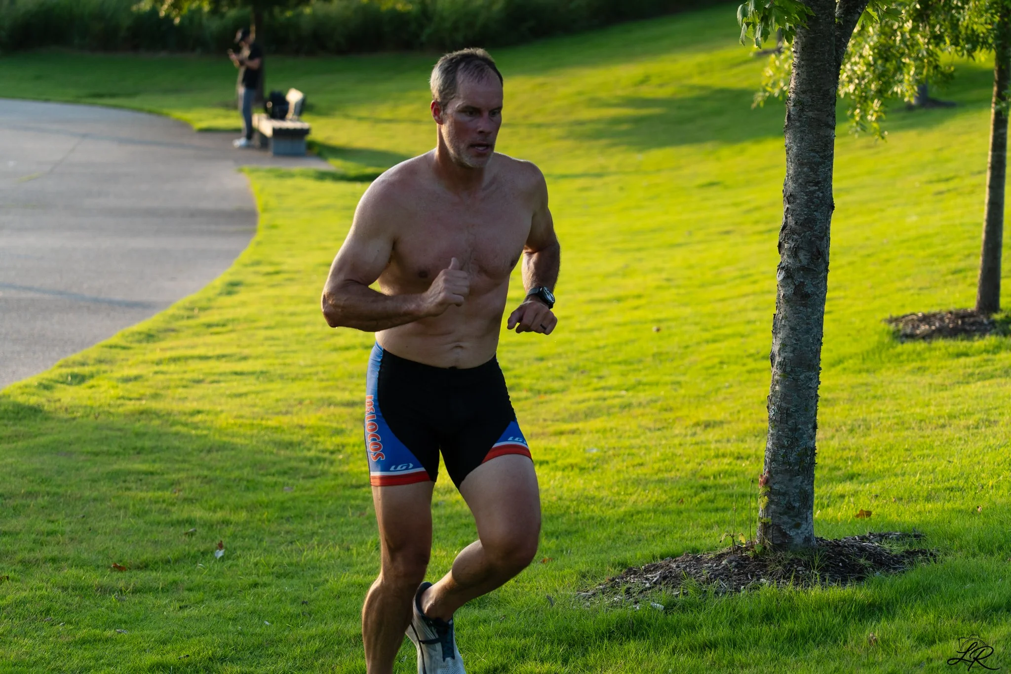 A middle-aged man running shirtless outdoors on a grassy path, wearing black and blue shorts, in a park during late afternoon or early evening, with trees and another person in the background.