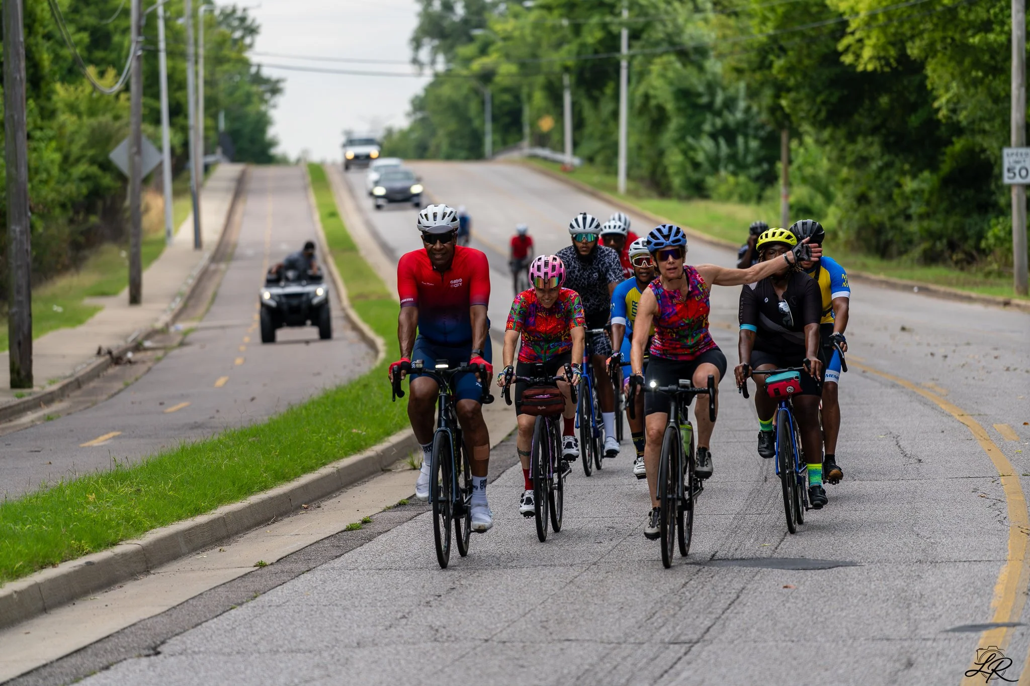 A group of cyclists riding on a road with cars passing by in the background, surrounded by green trees.