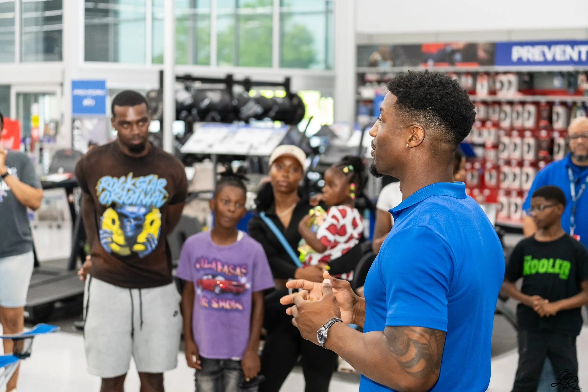 A man in a blue shirt is speaking to a group of diverse children and adults inside a store, with shelves of products in the background.
