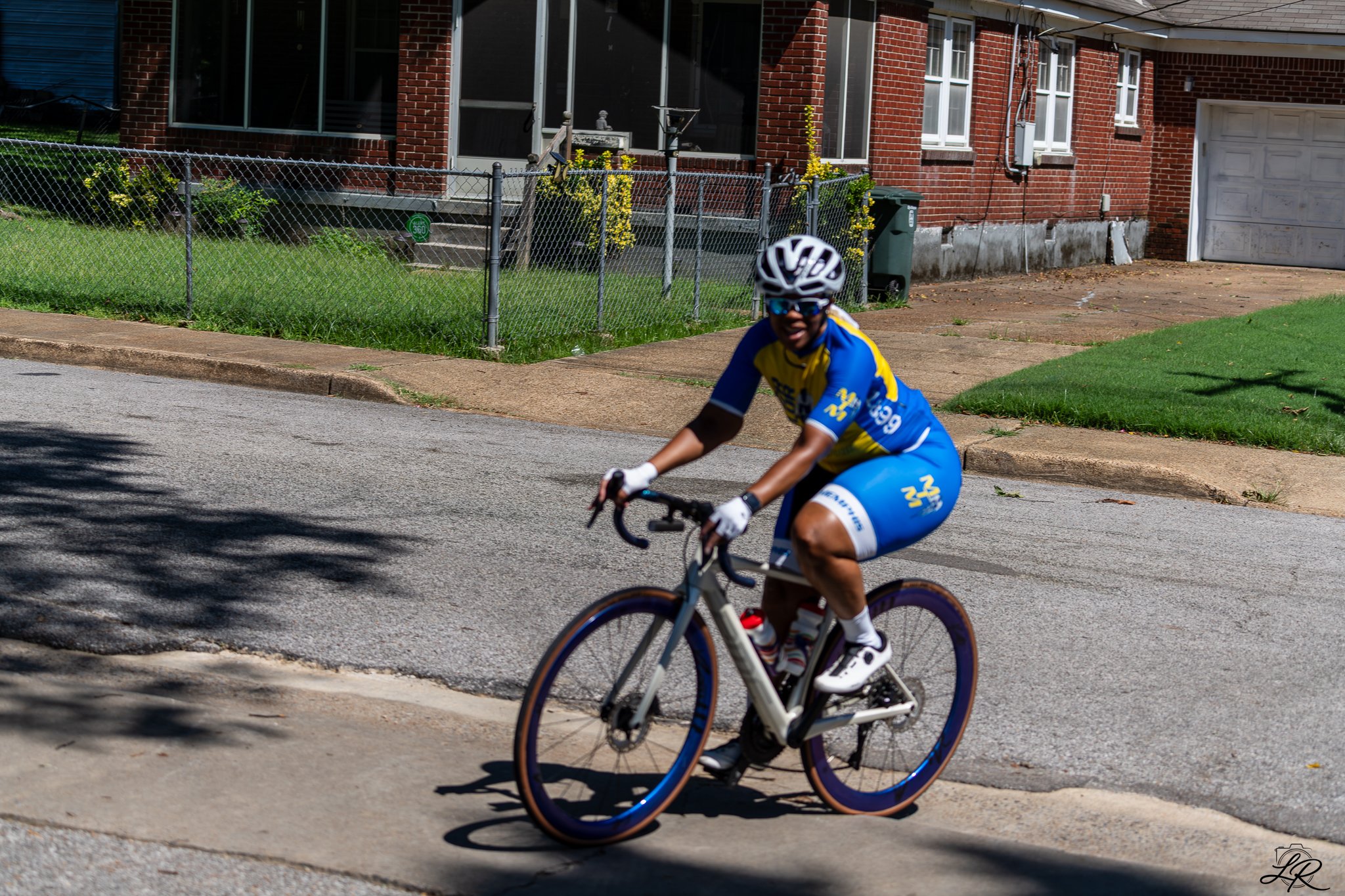 A person riding a bicycle on a residential street, wearing a helmet and sunglasses, with a brick house and a green trash bin in the background.