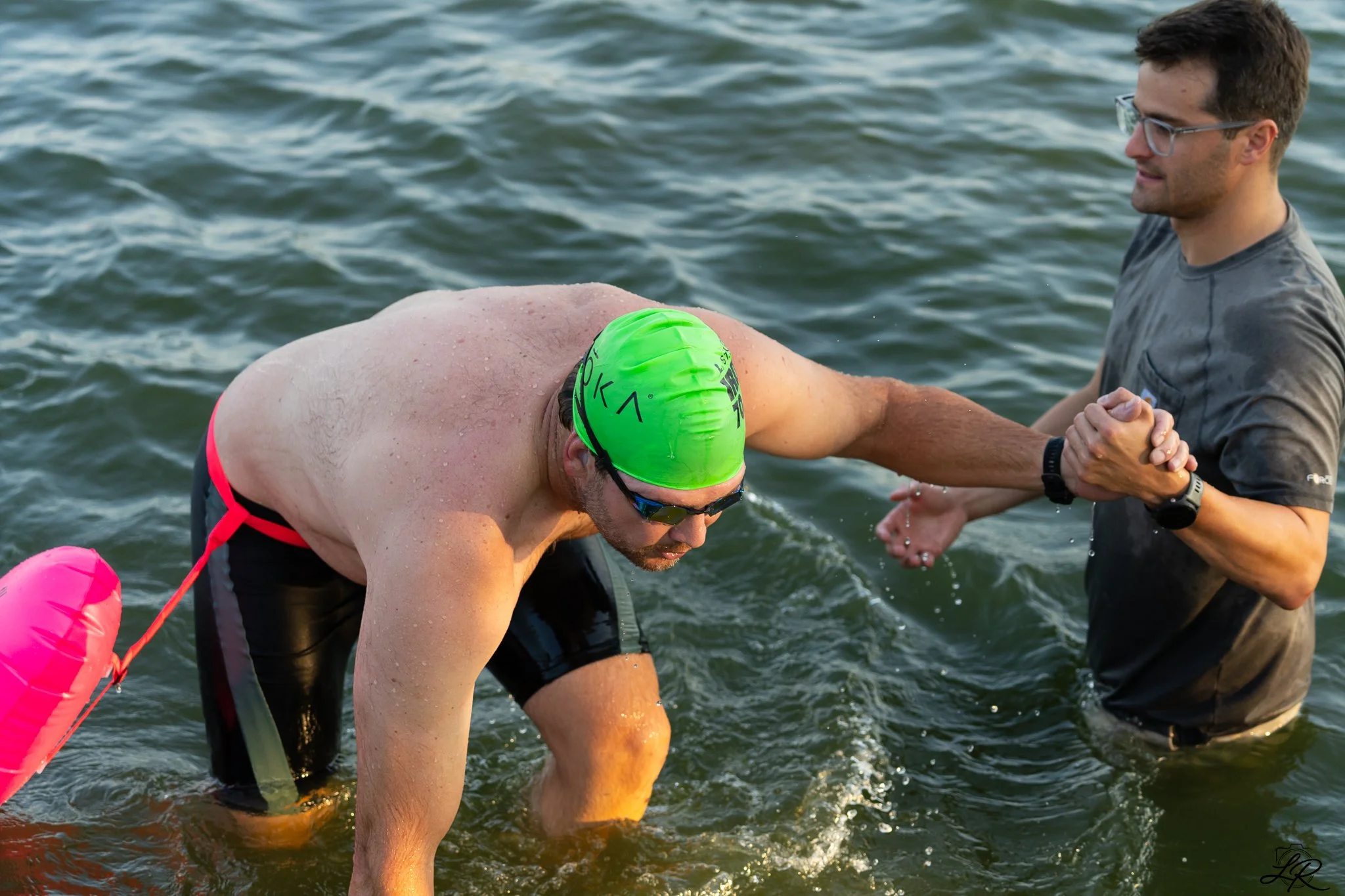 A man wearing a green swim cap and swimming goggles is helping another swimmer out of the water. The man is smiling and wearing a grey t-shirt, while the swimmer is bent over, with water droplets on his skin. They are in a body of water, likely a lak