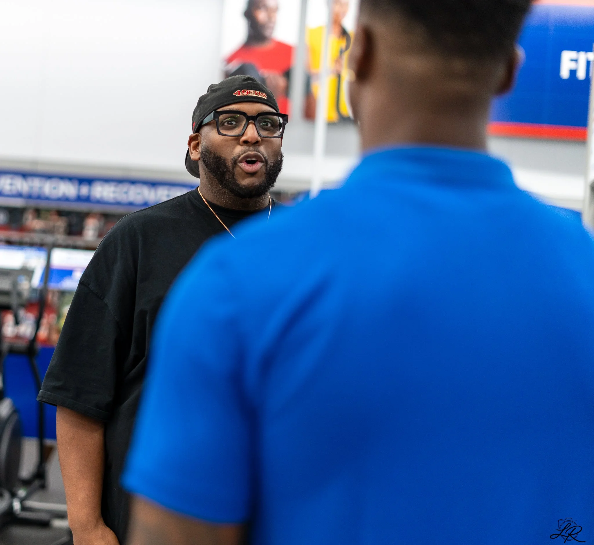 A man wearing glasses and a black beanie with red text, speaking to a person in a blue shirt in a store or sports venue. The background has a blurred poster and signage.