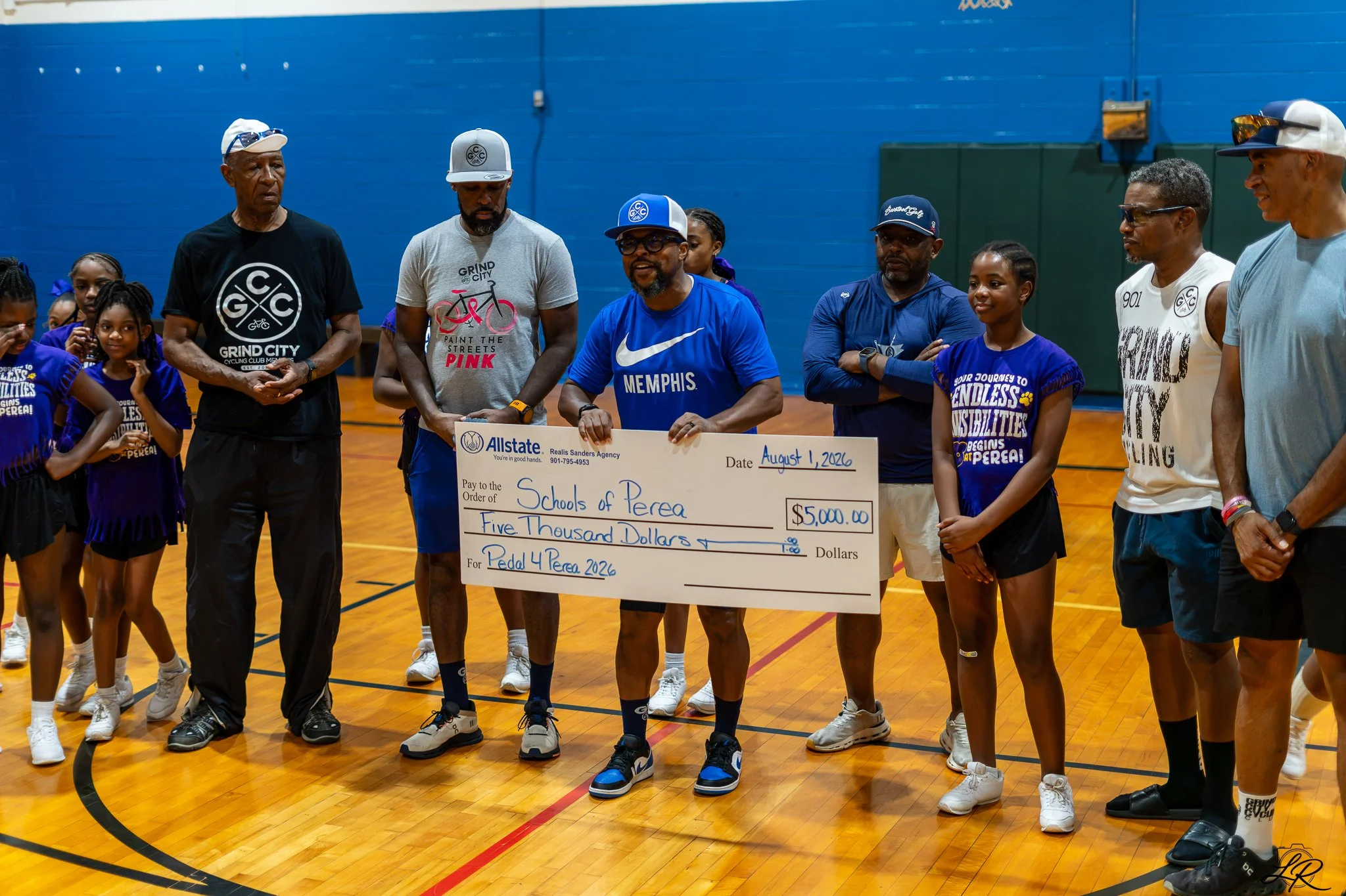 Group of people, including children and adults, standing on a gym floor during a presentation. They are holding a large check for five thousand dollars made out to Schools of Perea, dated August 1, 2026, from Allstate. Some people are smiling, and ot