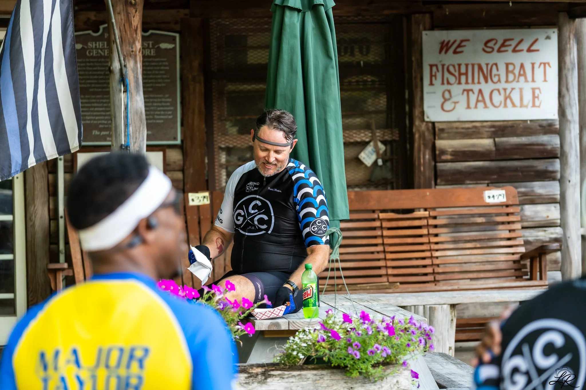 A man in cycling gear sitting at a table with a green Mountain Dew bottle in front of him, smiling and talking to another person with a blurred head in the foreground. The background has a sign that reads 'We Sell Fishing Bait & Tackle' and a wooden 
