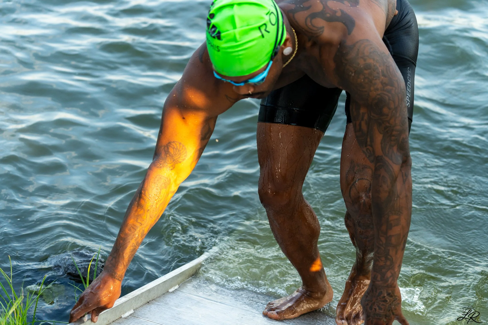 An athlete preparing to start a race on a dock by the water, wearing a bright green swim cap, goggles, and black athletic gear, with tattoos on their arms and legs.