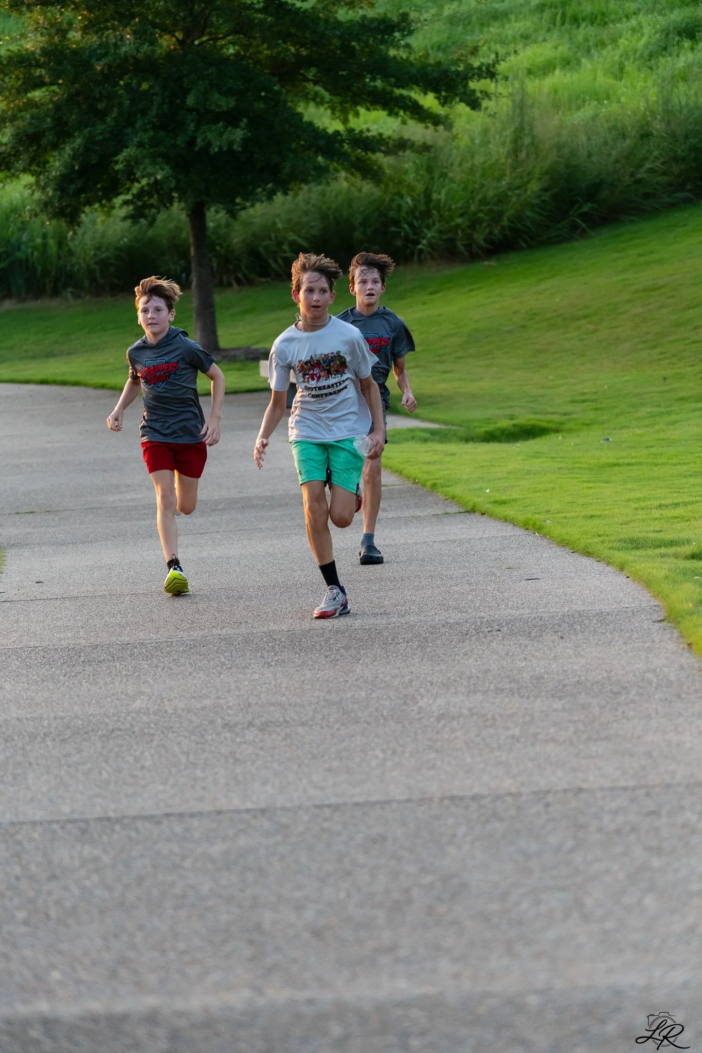 Three young boys running on a paved trail in a park with trees and grass in the background.